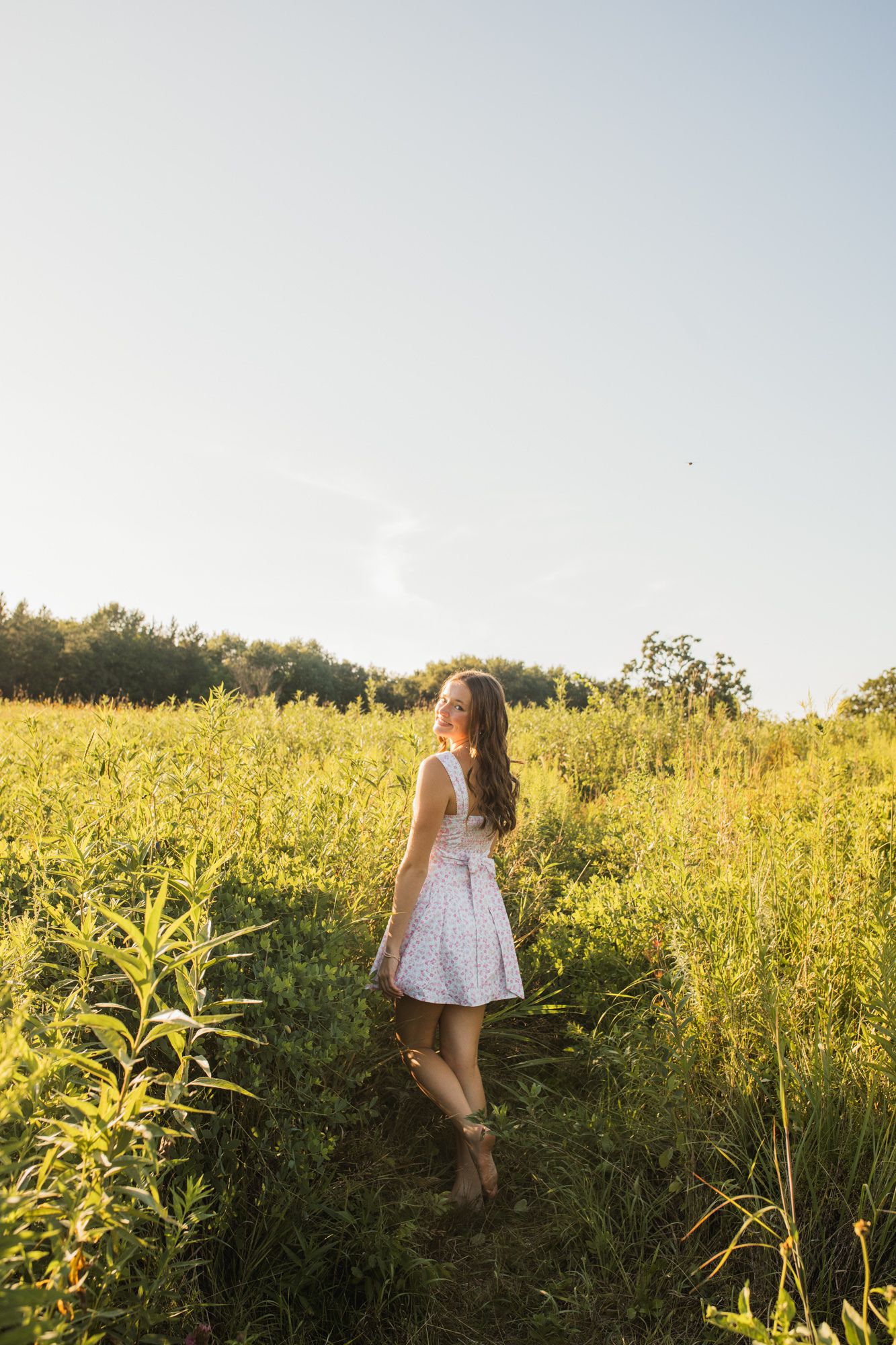 Young woman in white dress walking through a sunlit green field on a warm summer day.
