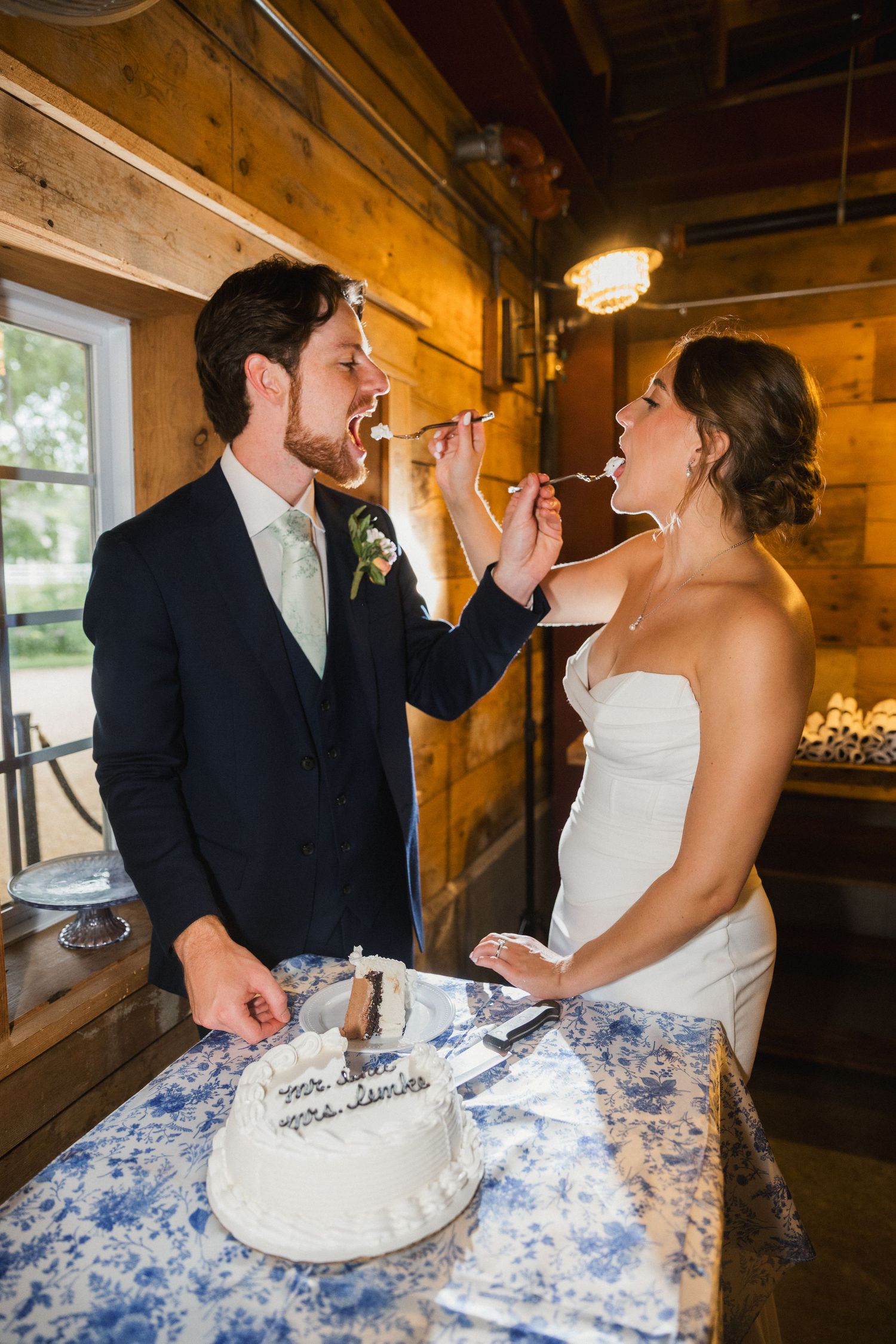Bride and groom feeding each other wedding cake at rustic barn reception venue.