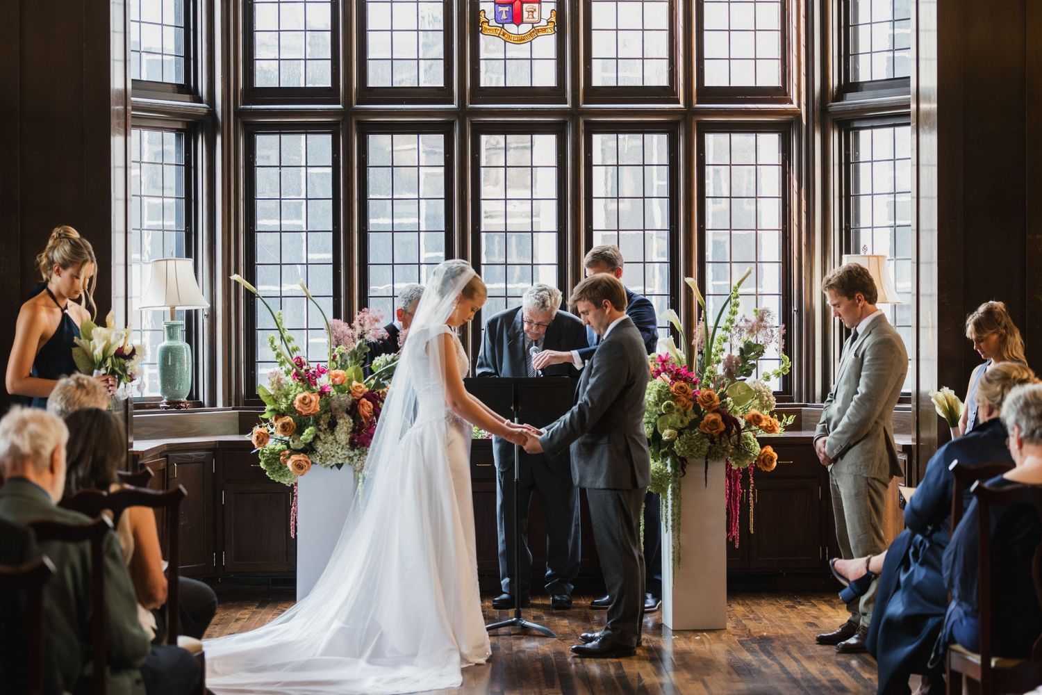 Wedding ceremony with bride and groom holding hands before officiant, framed by large windows.