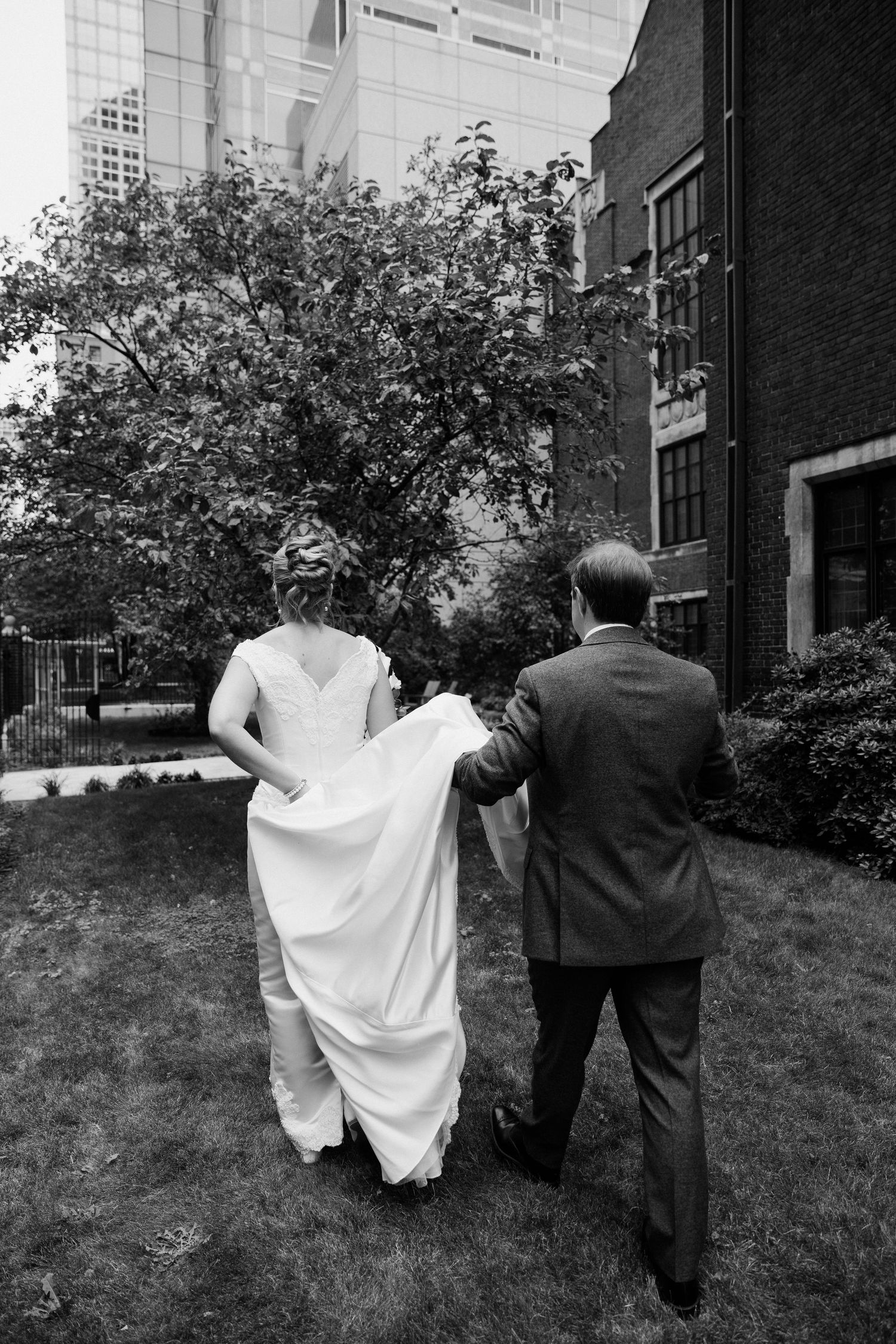 Bride and groom walking outdoors, man carrying wedding dress train, black and white photo.