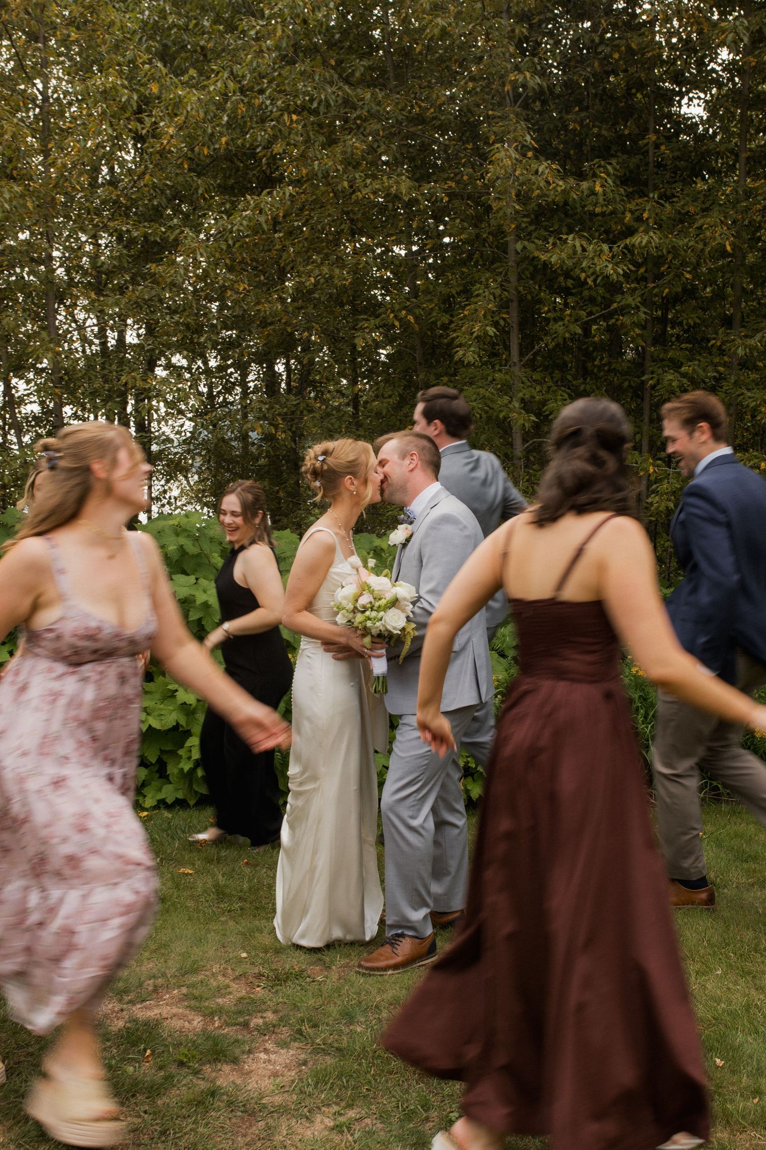 Bride and groom share a kiss outdoors surrounded by wedding guests in formal attire near lush green trees.