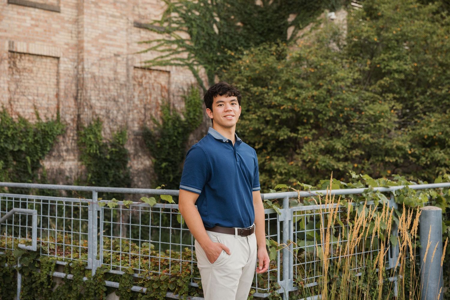 Young man in blue polo shirt and khaki pants standing by metal railing with lush greenery and brick building behind him.
