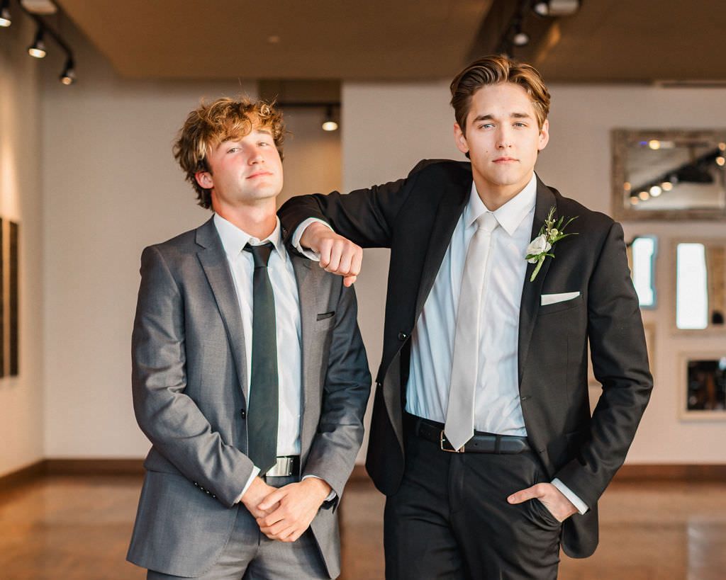 Two groomsmen in suits pose casually in wedding venue hallway.
