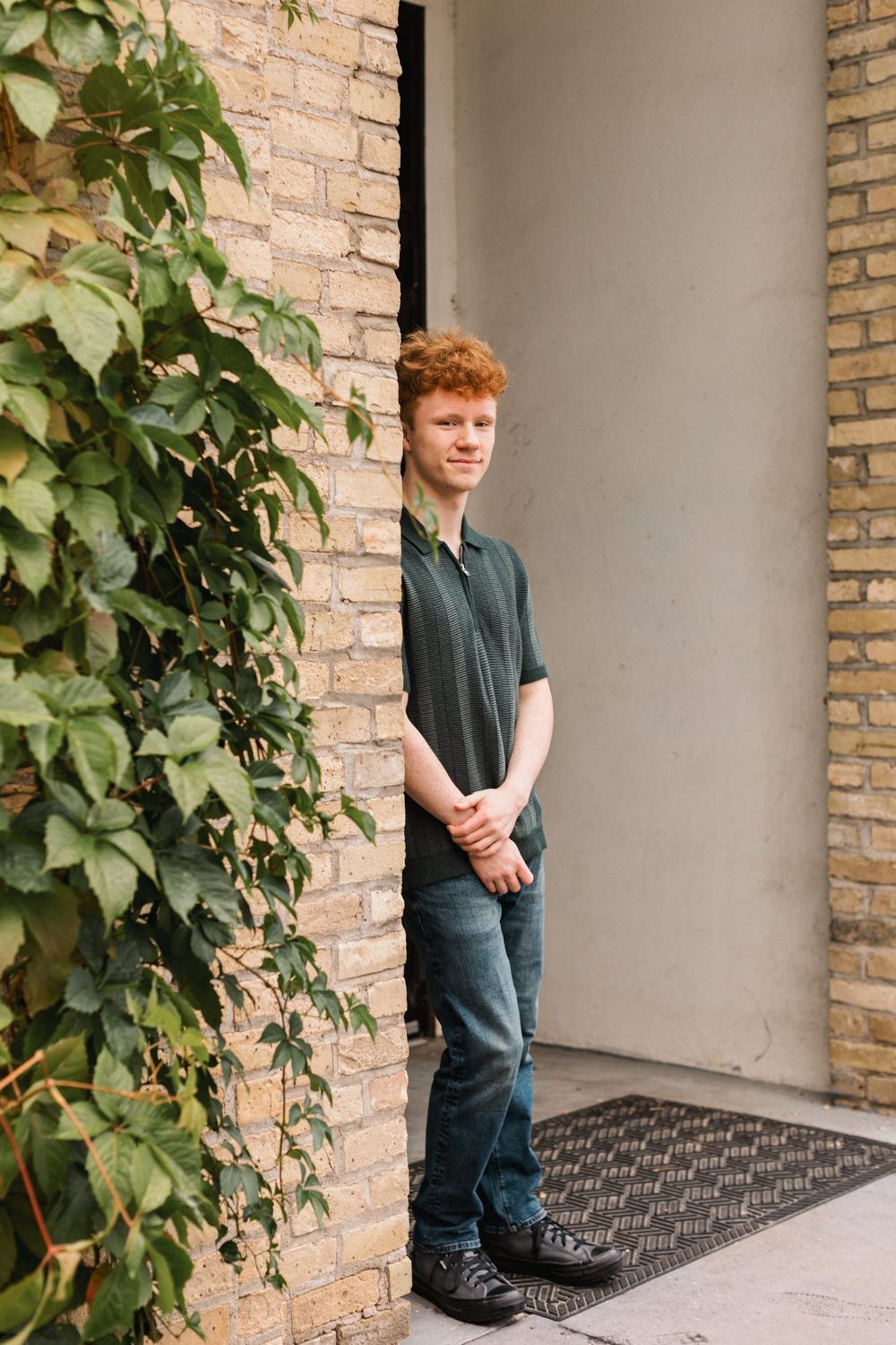 A person posing in a casual outfit near a brick wall with green foliage and architectural columns.