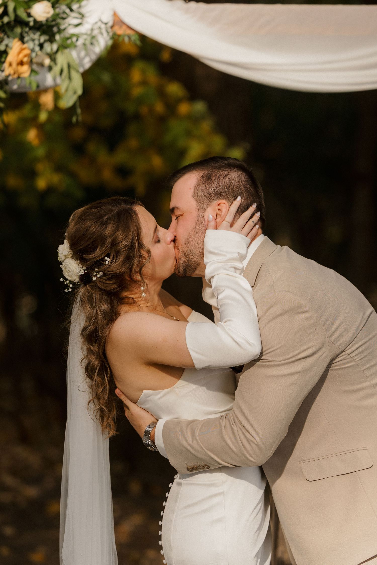 Bride and groom share a wedding ceremony kiss under a draped floral arch surrounded by golden autumn foliage.