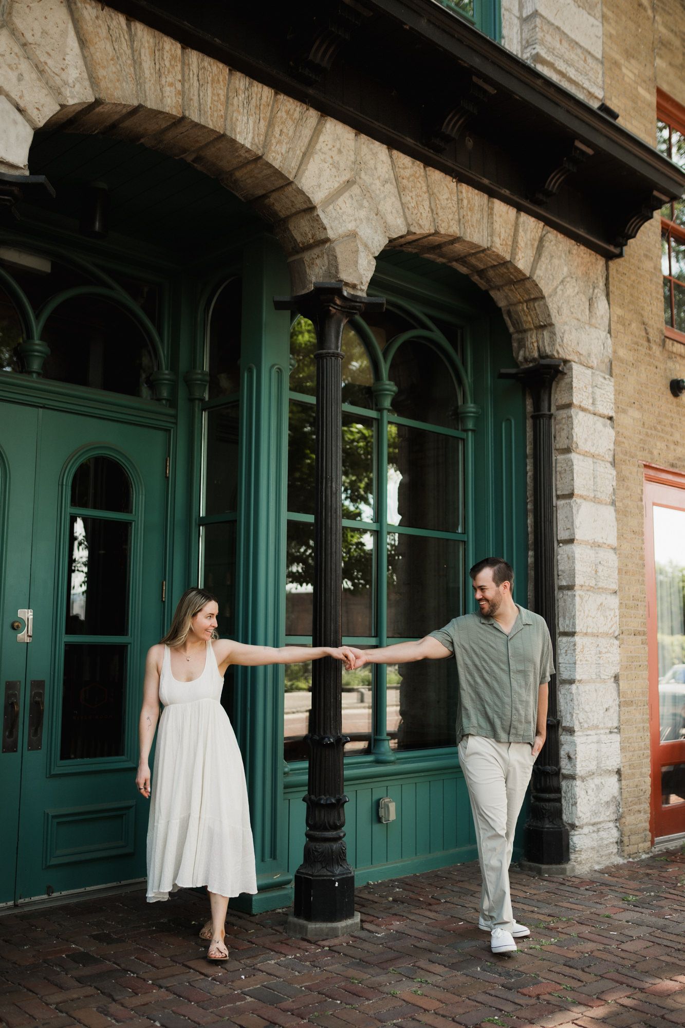 Couple holds hands around a black lamppost outside a building with green arched doors and stone facade.