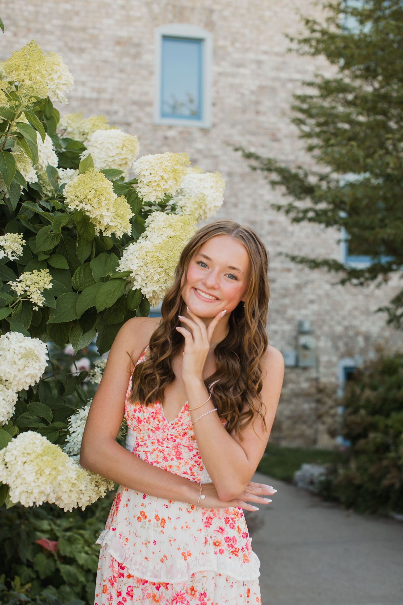 Smiling teenage girl in floral dress posing beside white hydrangea blooms near a stone building.