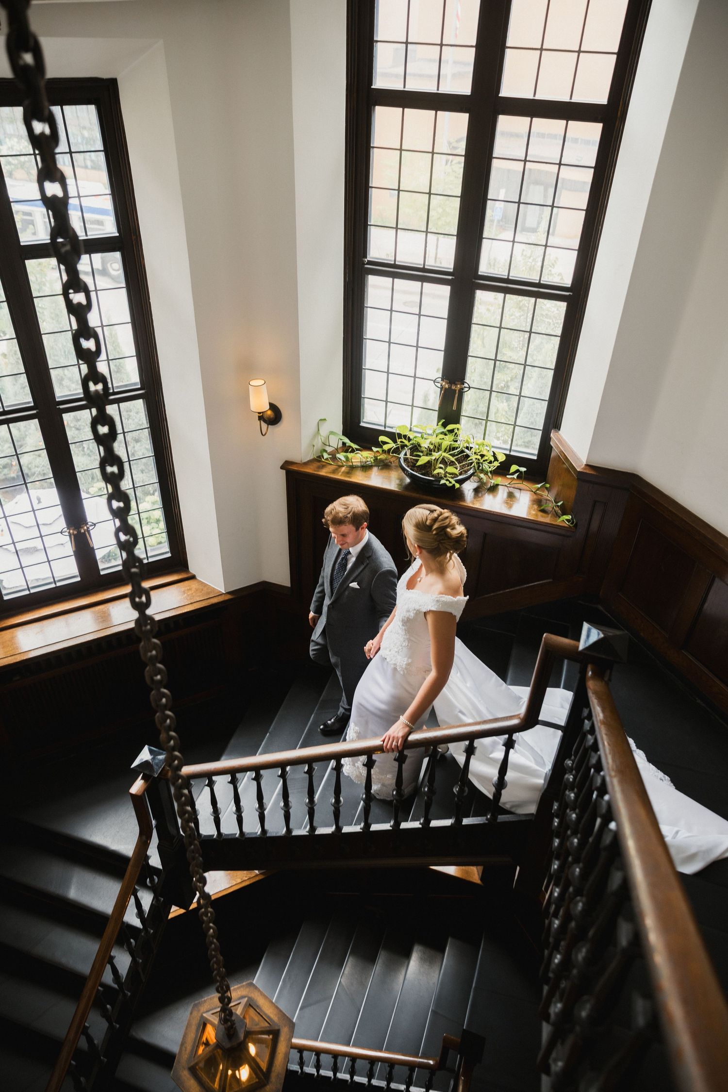Aerial view of bride and groom walking up dark wooden staircase in elegant historic venue with tall windows.
