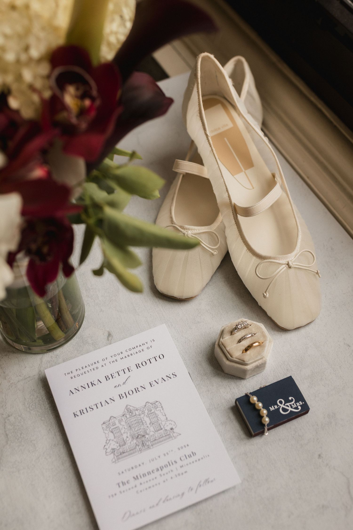 Elegant bridal flat shoes with bow detail beside wedding invitation, jewelry box, and floral bouquet on marble surface.