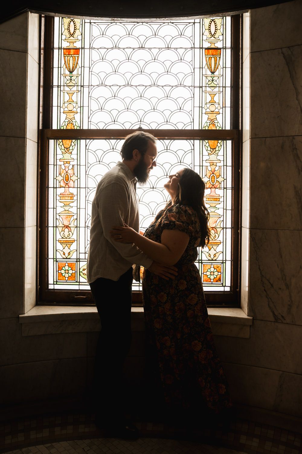 A romantic couple embraces in front of stained glass windows in a historic building.
