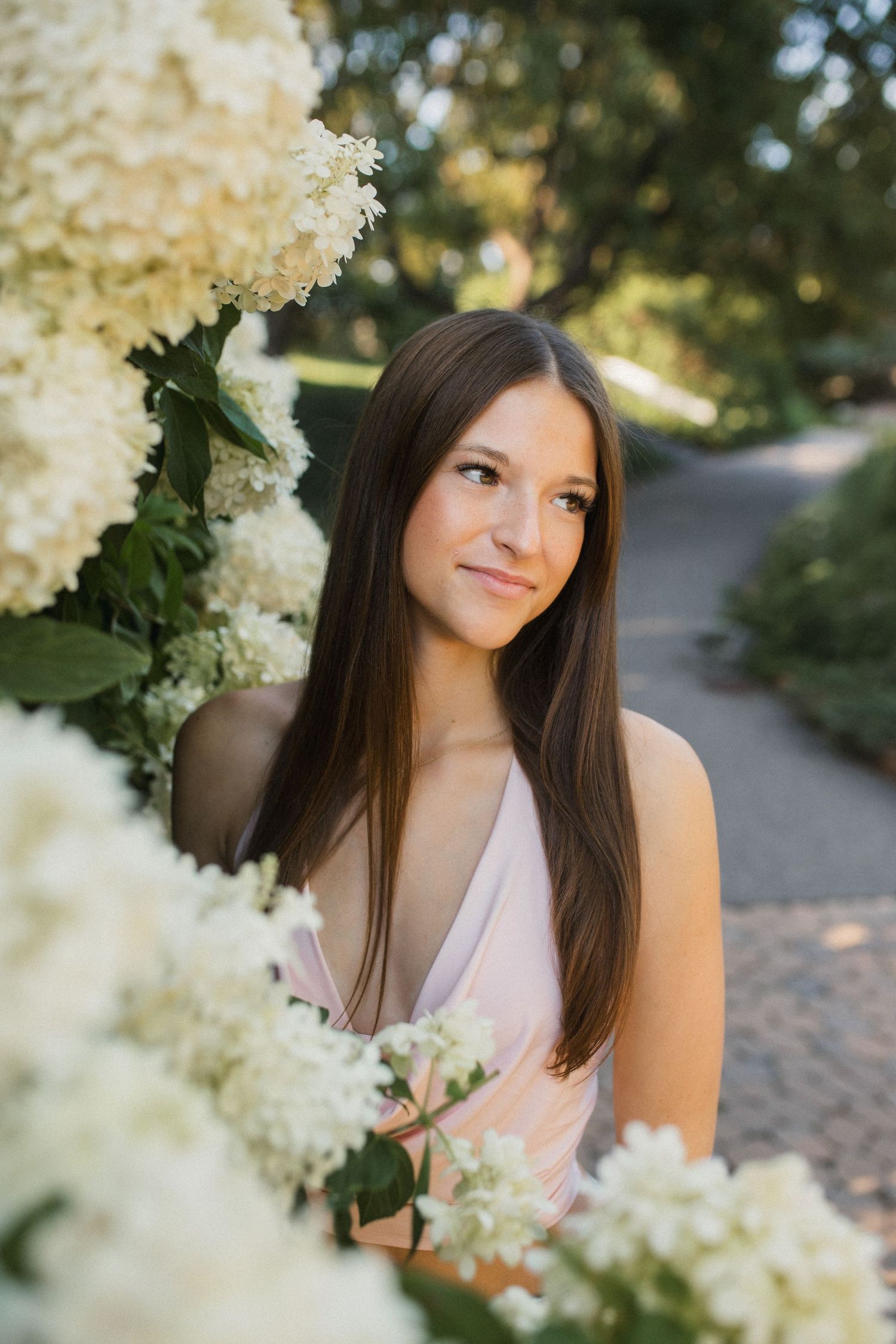 Young woman with long brown hair in pink dress surrounded by white hydrangea blooms in a garden setting.