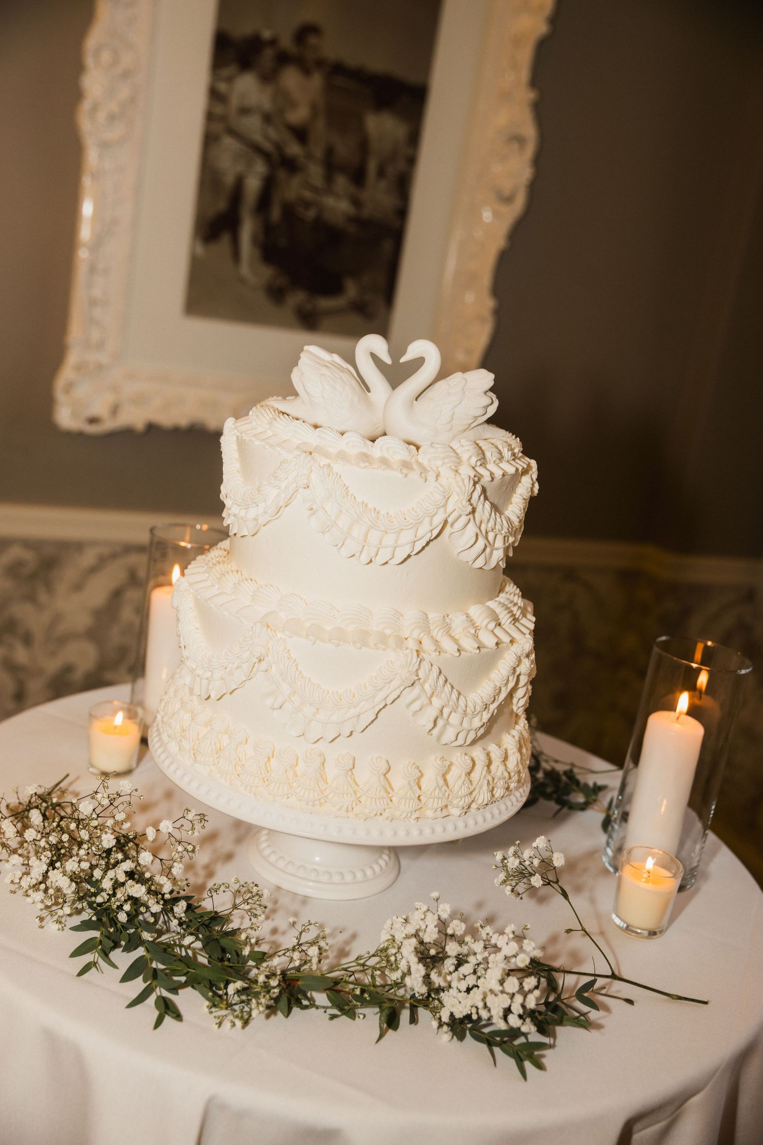 Elegant three-tier white wedding cake with swan topper, piped frosting details, candles, and baby's breath floral arrangement.