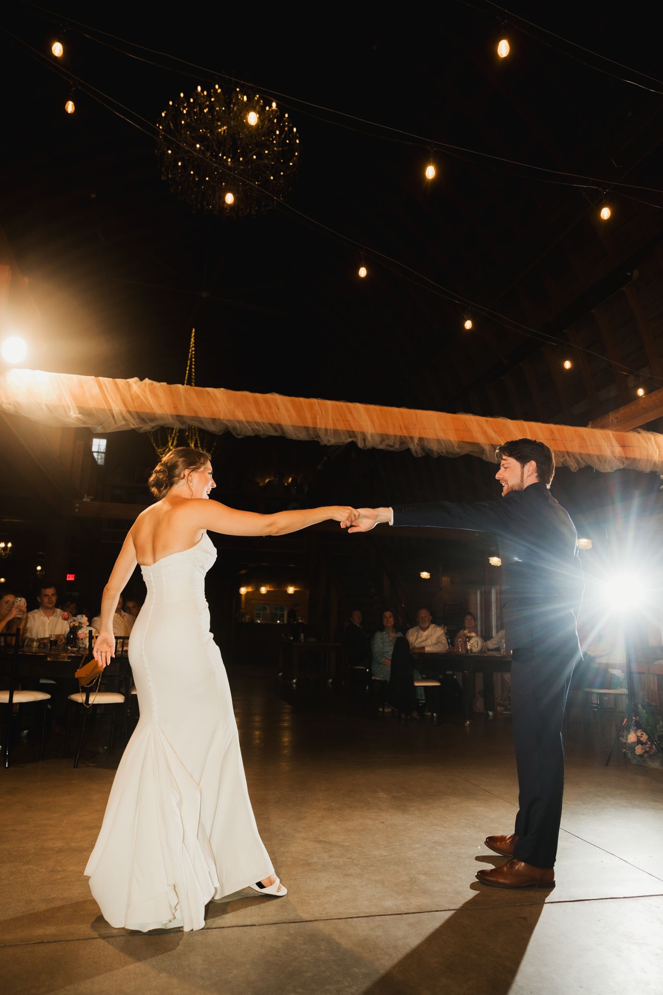 Bride and groom share their first dance under warm string lights and chandelier at elegant wedding reception.