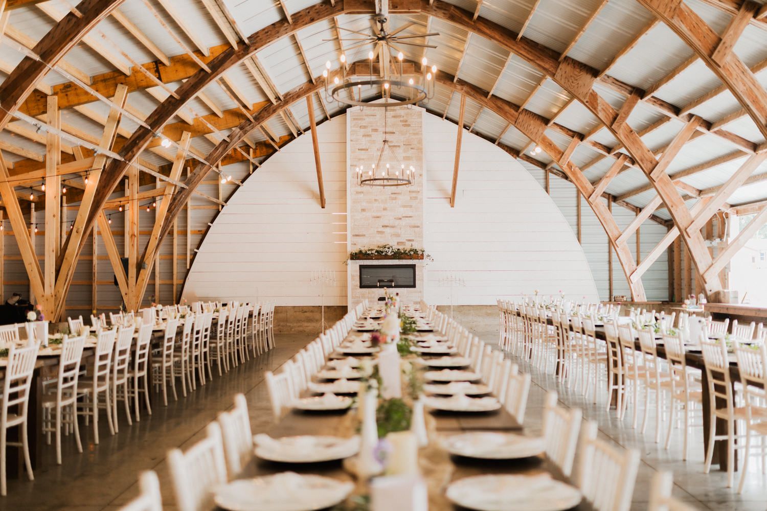 Long rustic reception hall with exposed wooden beams and white walls set for an elegant wedding dinner.