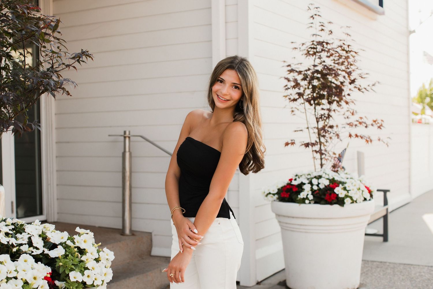 Young woman in black strapless top and white pants poses outdoors near white flower planters and building steps.