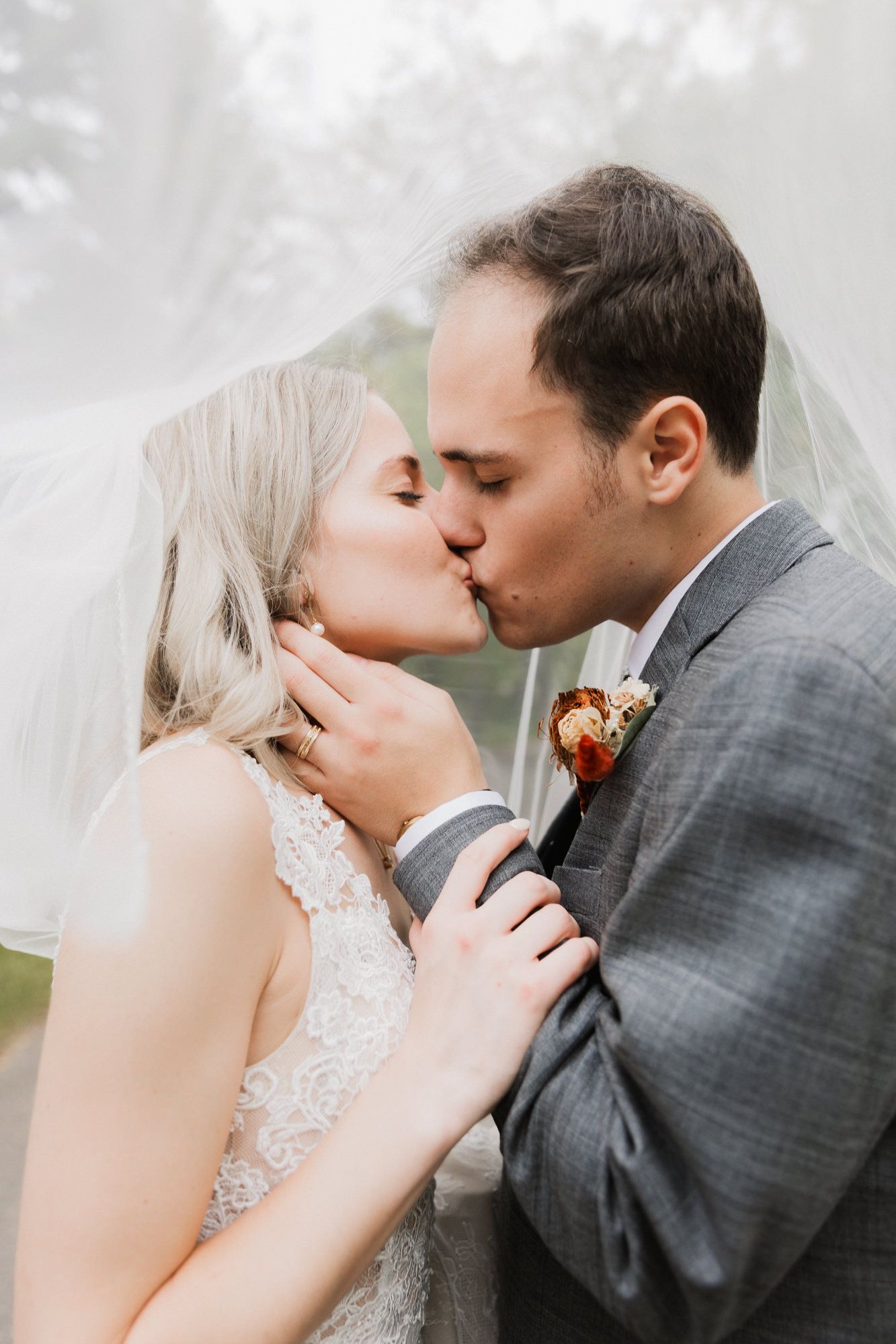 Bride and groom share a romantic kiss under a flowing veil on their wedding day, with the groom wearing a gray suit.