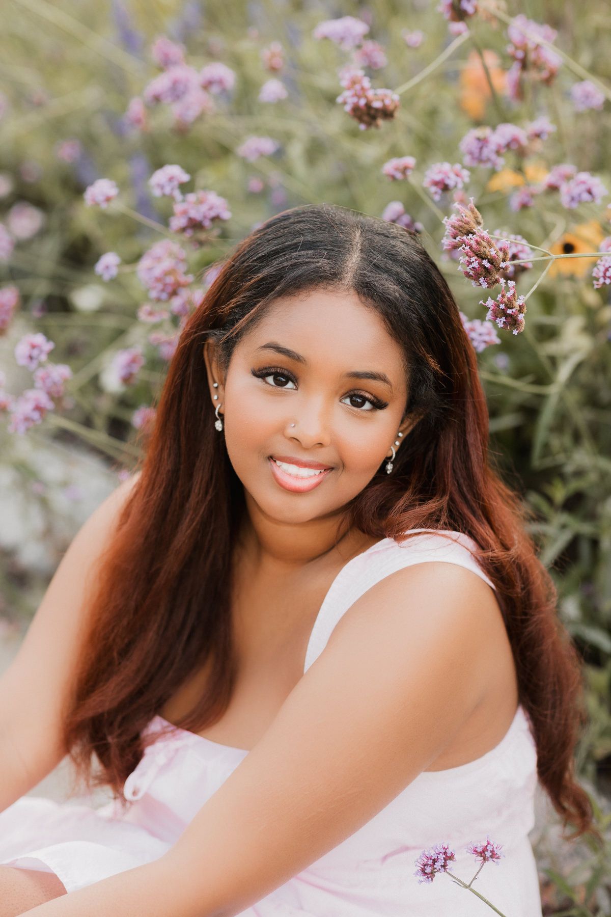 Young woman in white dress smiling outdoors among purple wildflowers, with long auburn hair and drop earrings.