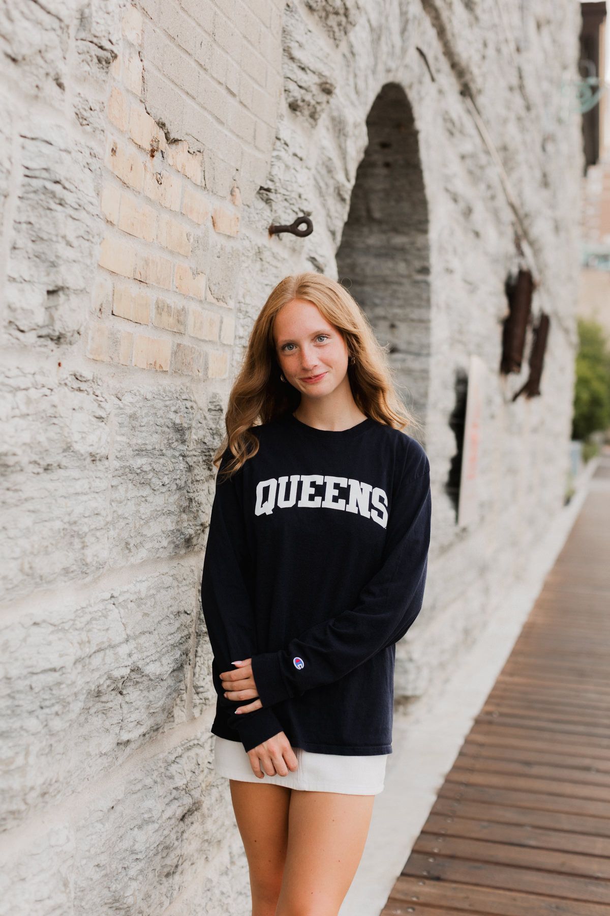 Teen girl in Queens Champion sweatshirt posing against white brick wall on wooden walkway.