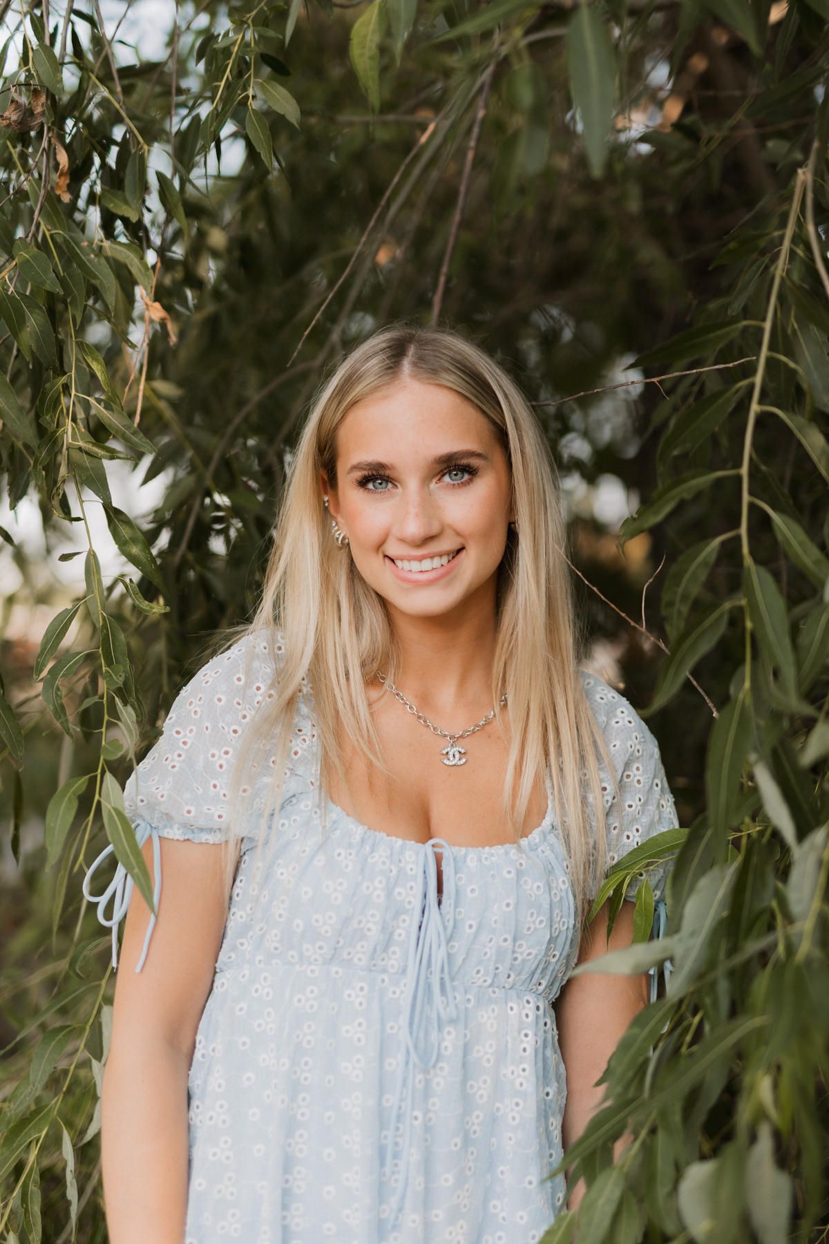 Smiling blonde girl in light blue eyelet dress standing among weeping willow branches outdoors.