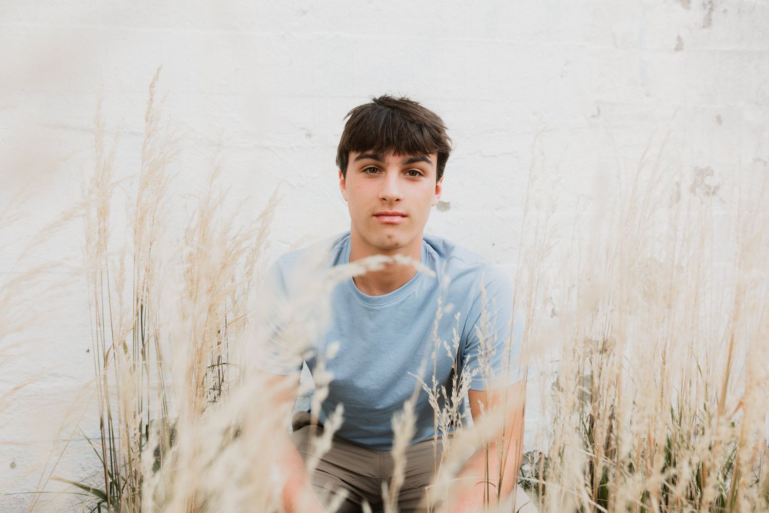 Young man in light blue shirt sitting among tall dry grasses against a white wall background.
