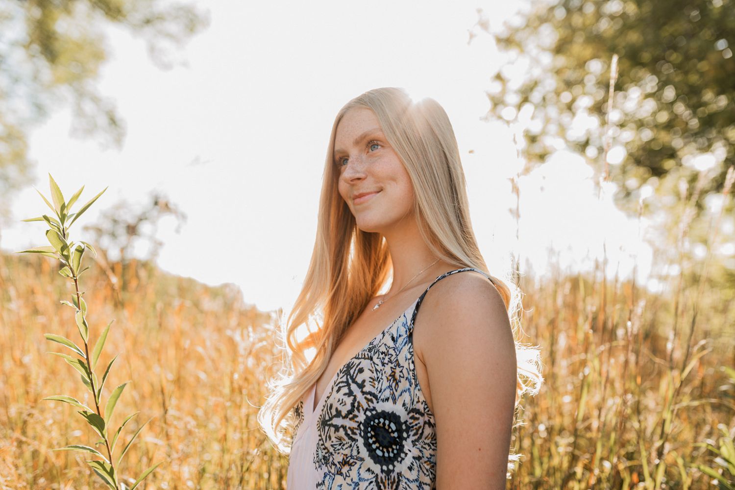 Blonde teenage girl in floral sundress smiling softly in a golden sunlit field of tall grass.