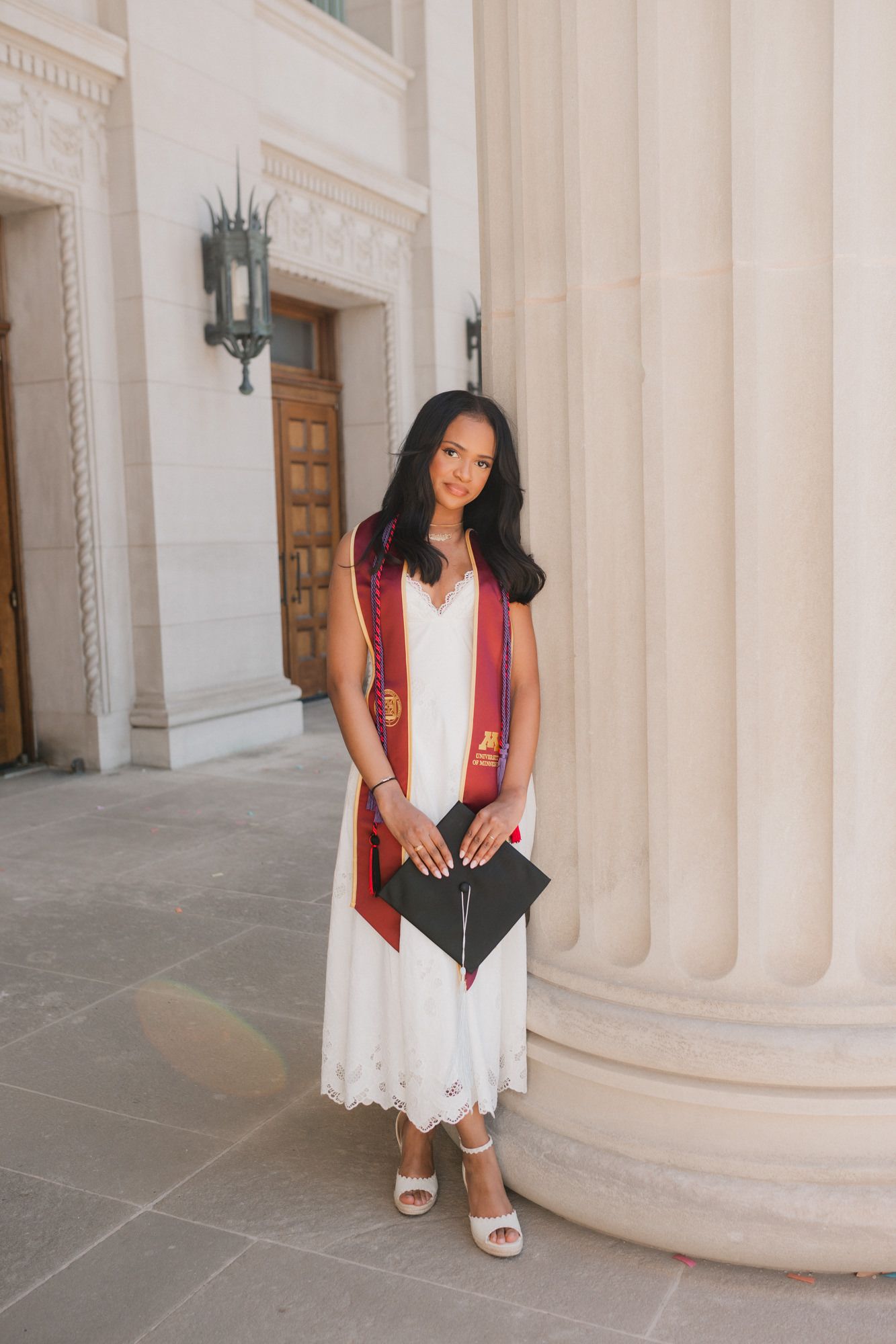 A young woman in a white dress and red graduation stole holds her cap outside a classical stone building.