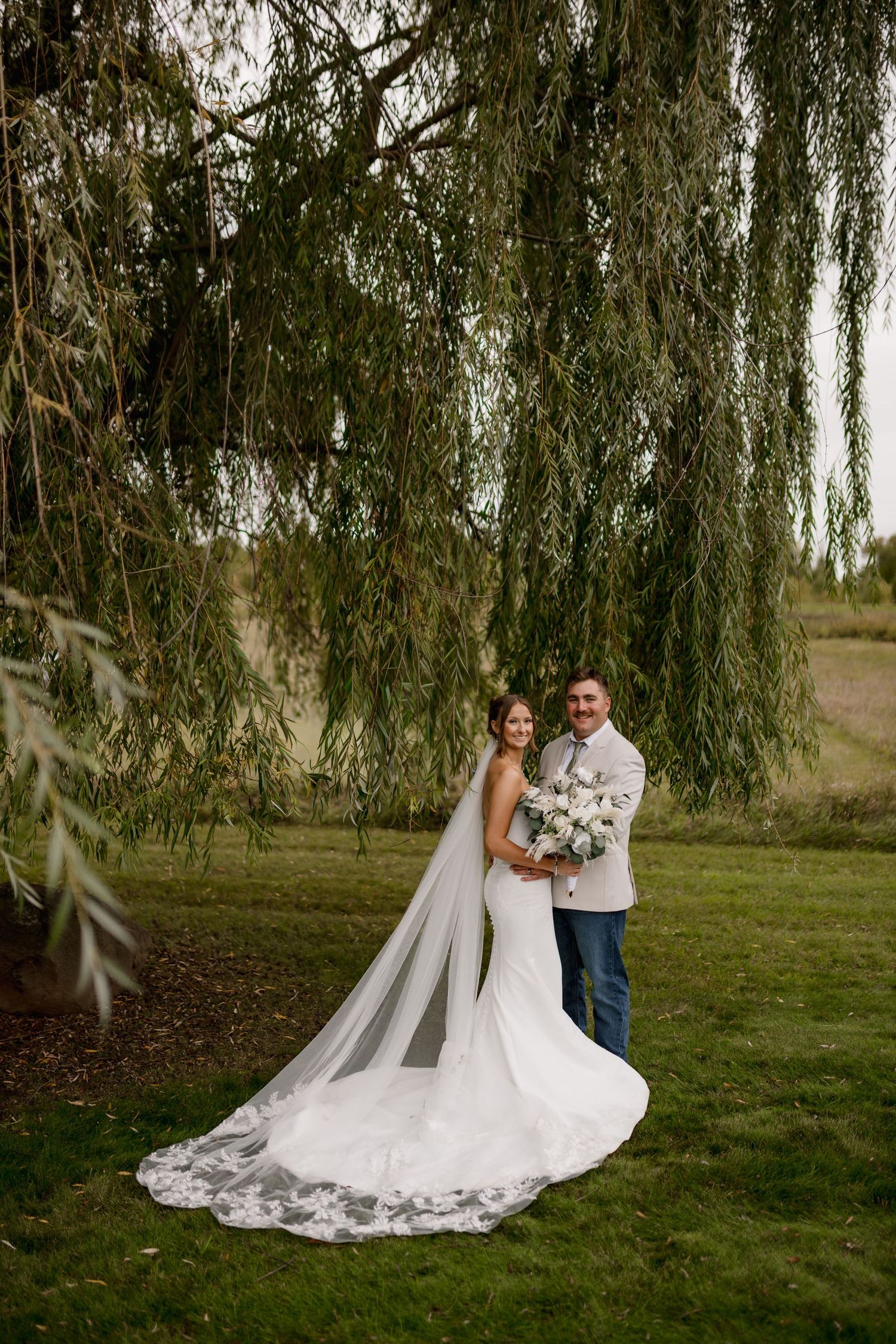 Bride and groom posing under weeping willow tree, bride in fitted lace gown with cathedral veil.