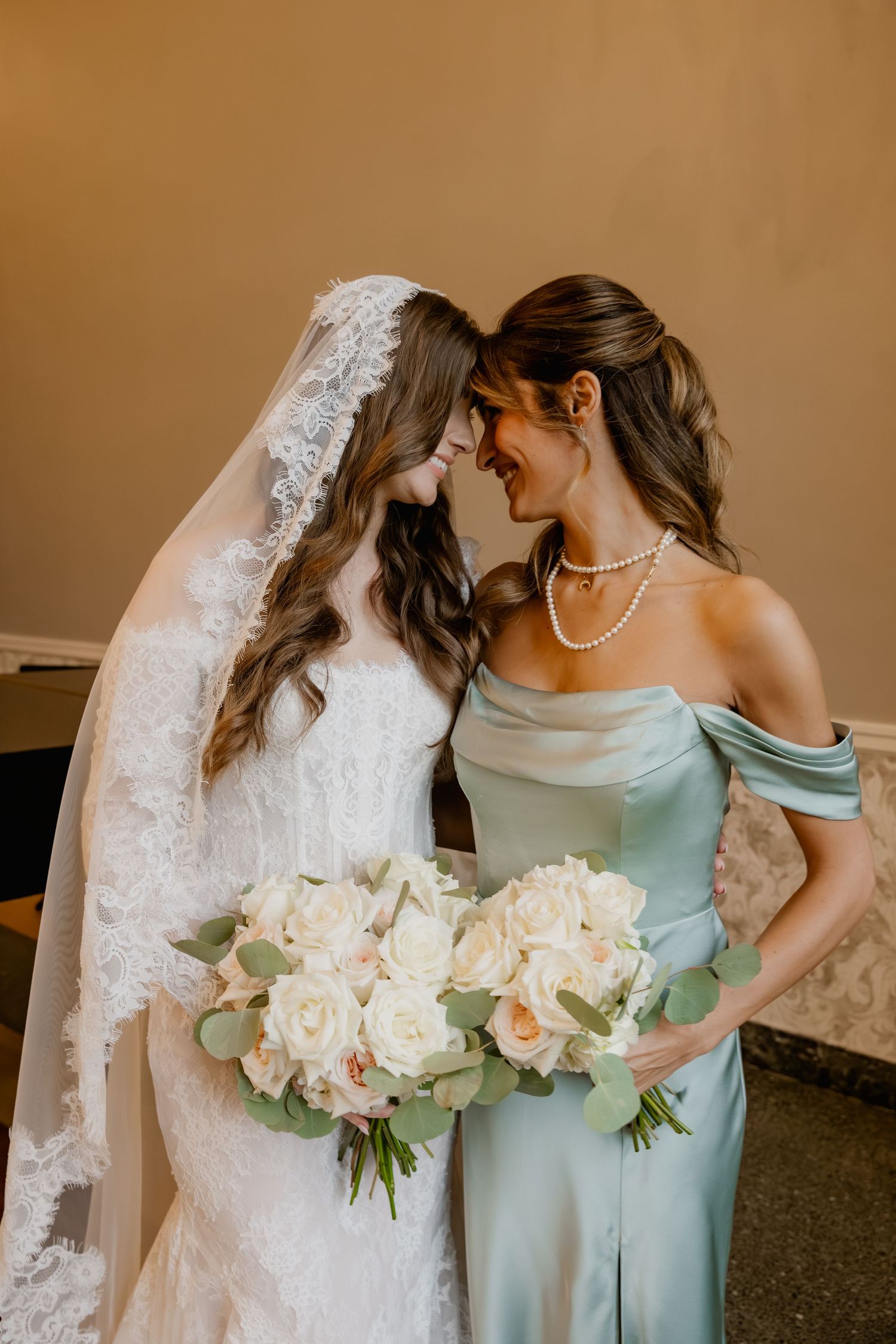 Bride in lace gown and veil touching foreheads with bridesmaid in sage off-shoulder dress, holding white rose bouquet.