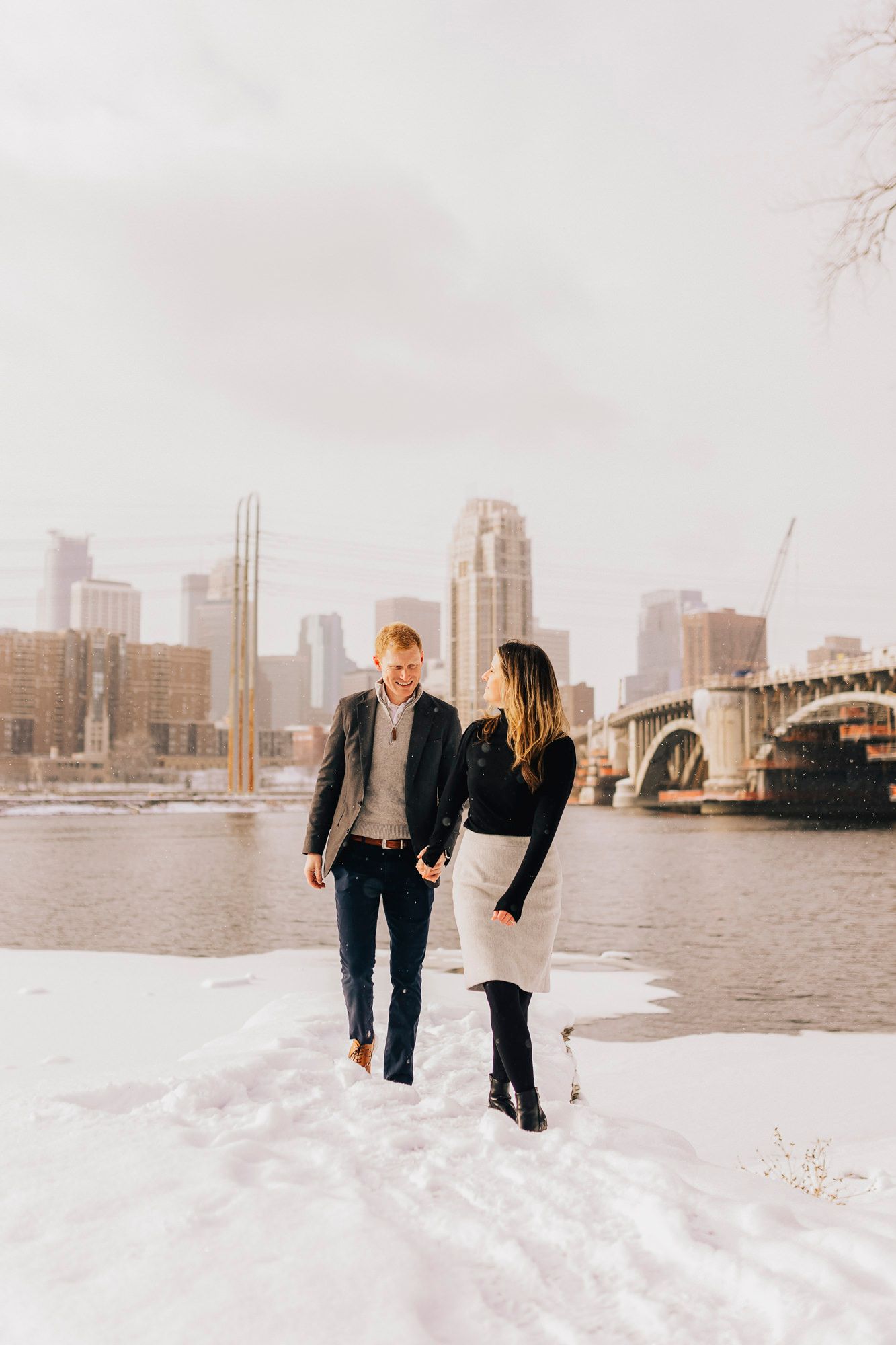 A romantic winter engagement photo with a city skyline backdrop and snow covered ground.