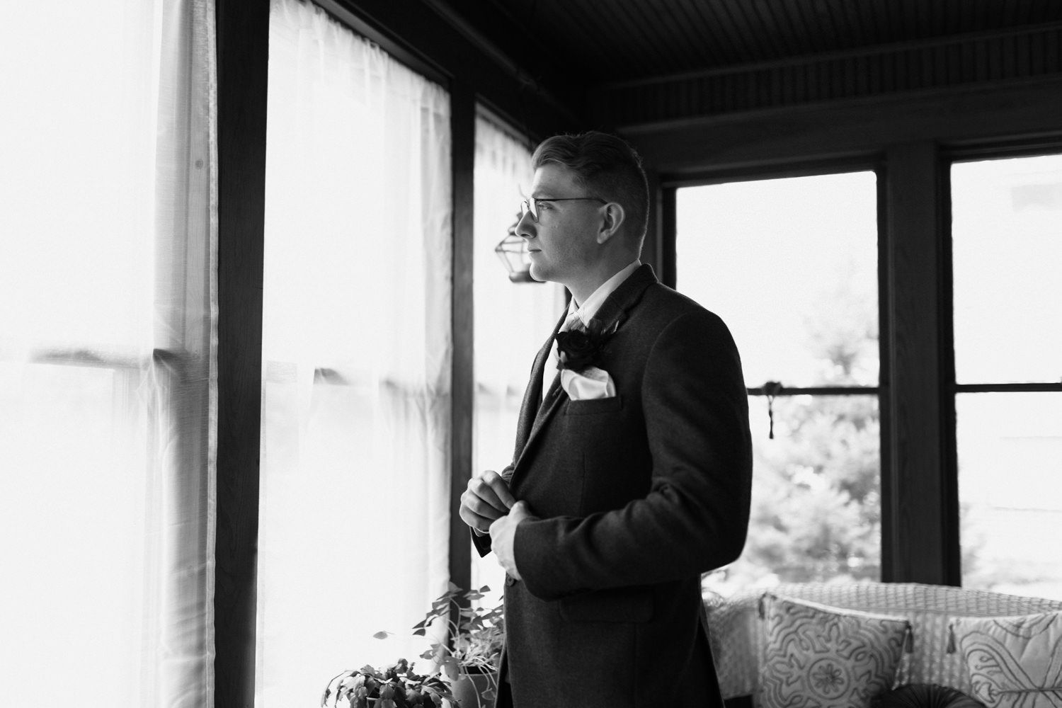 Black and white photo of a groom looking out window in a rustic enclosed porch.