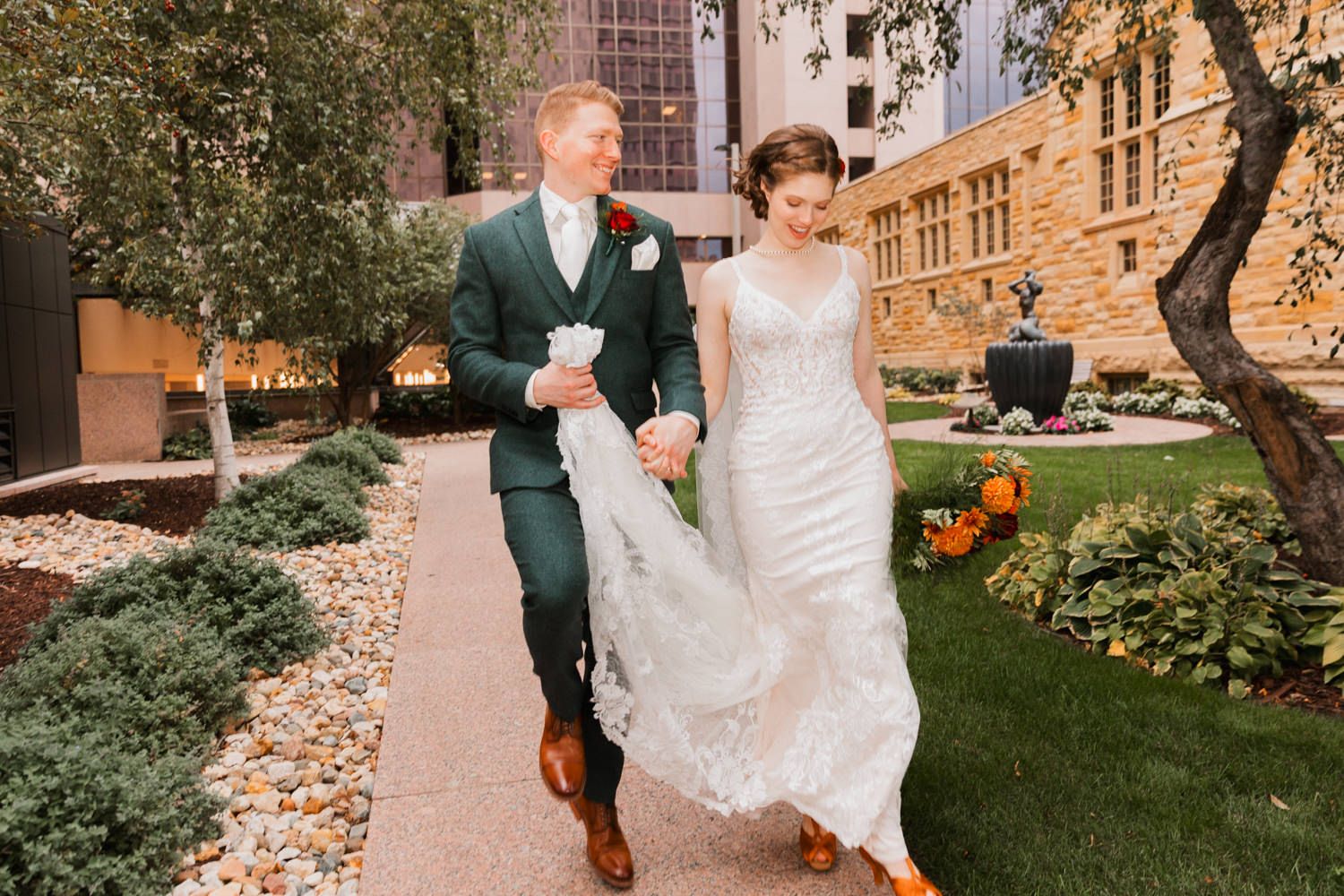 A couple walks together on a garden path outside a historic stone building on their wedding day.