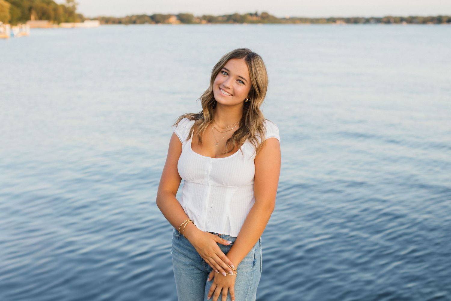 Young woman in white top and jeans smiling by a serene lake at golden hour.