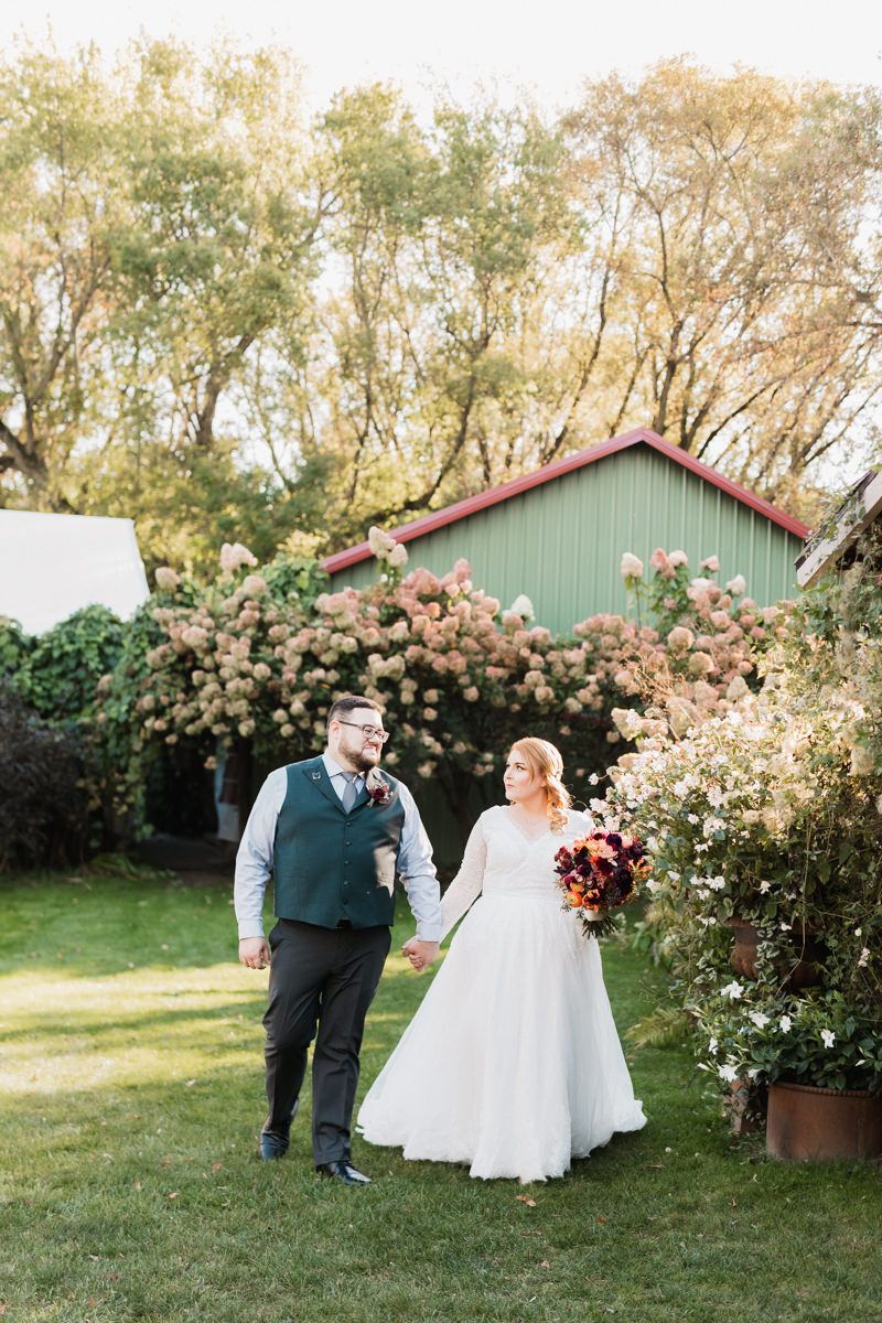 Wedding couple poses for portraits in front of a rustic green barn surrounded by flowering rose bushes.