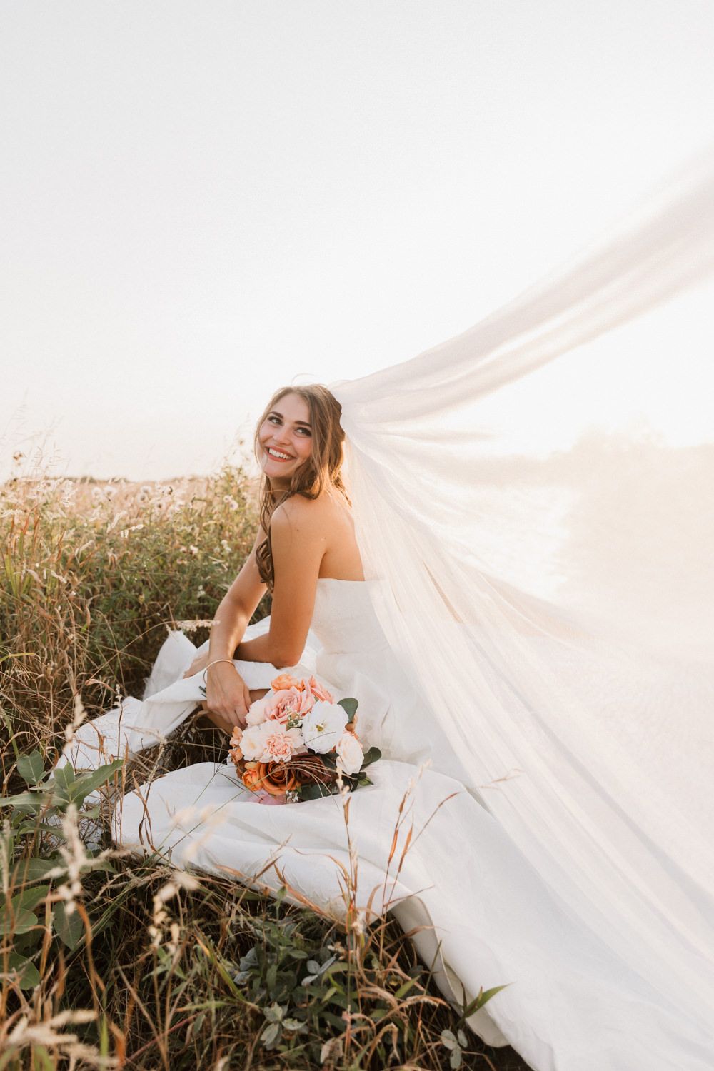 Bride in white dress poses with veil flowing in golden sunlight during outdoor photo session.