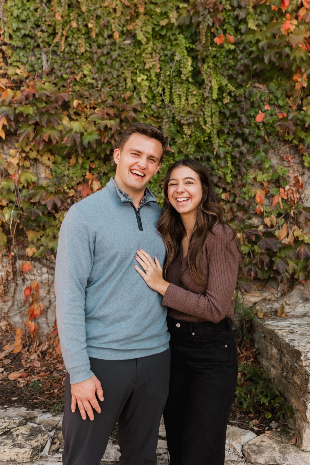A couple shares a joyful moment together against a backdrop of autumn foliage and stone wall.