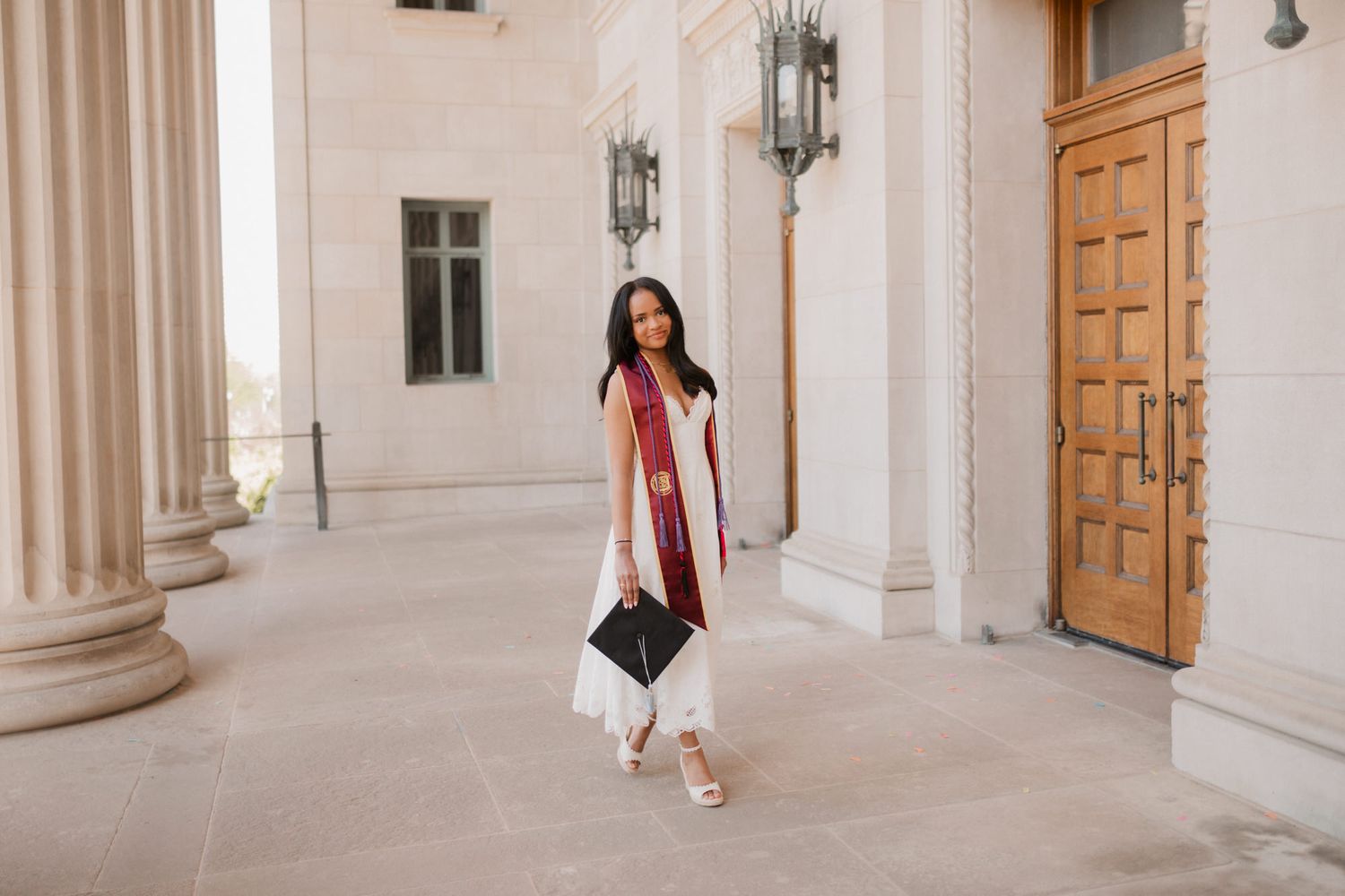 Young woman in white dress with maroon graduation stole holds cap while walking near ornate building entrance.