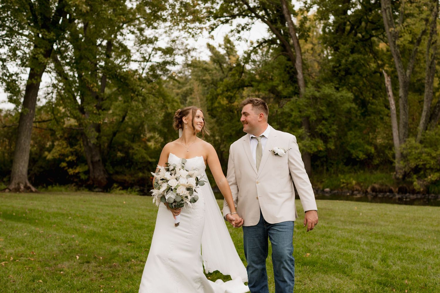 Bride and groom walk hand-in-hand on green lawn, she holds white bouquet, he wears tan blazer with jeans.