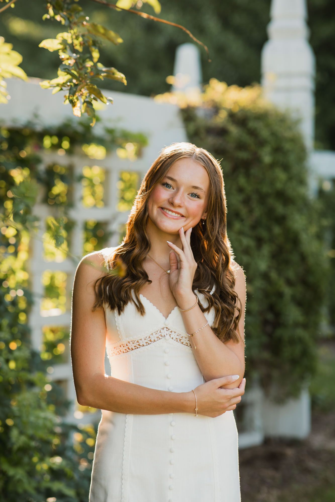 Young woman in white sundress smiling outdoors near white lattice with lush green foliage in warm golden sunlight.