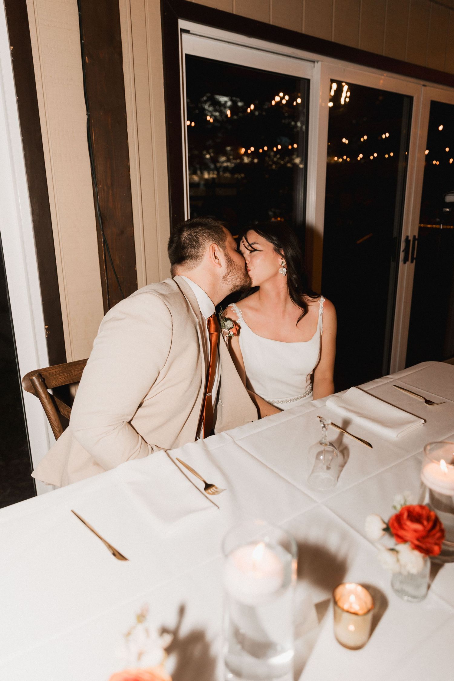Romantic couple shares intimate moment at candlelit dinner table with white tablecloth and rose centerpiece.
