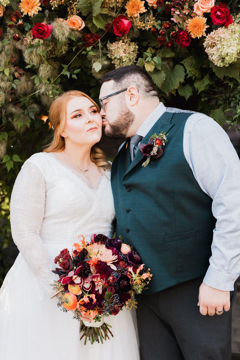 A wedding couple poses in front of a floral arch with orange, red and white blooms as the bride holds a burgundy bouquet.