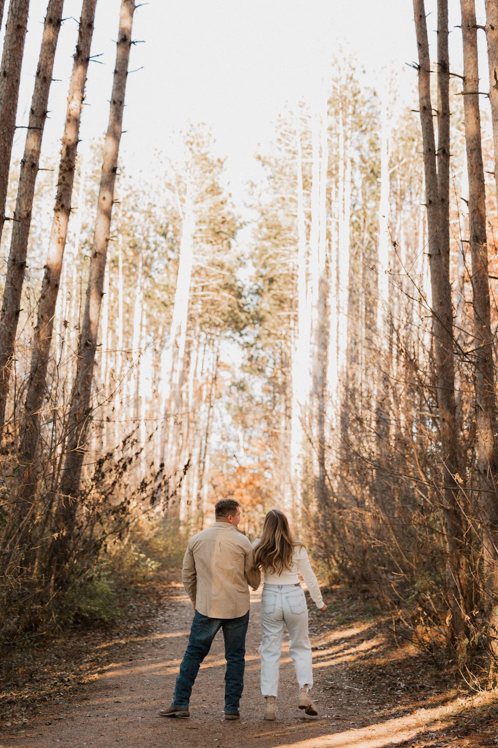 Two people stroll together along a wooded path surrounded by towering trees in golden afternoon light.