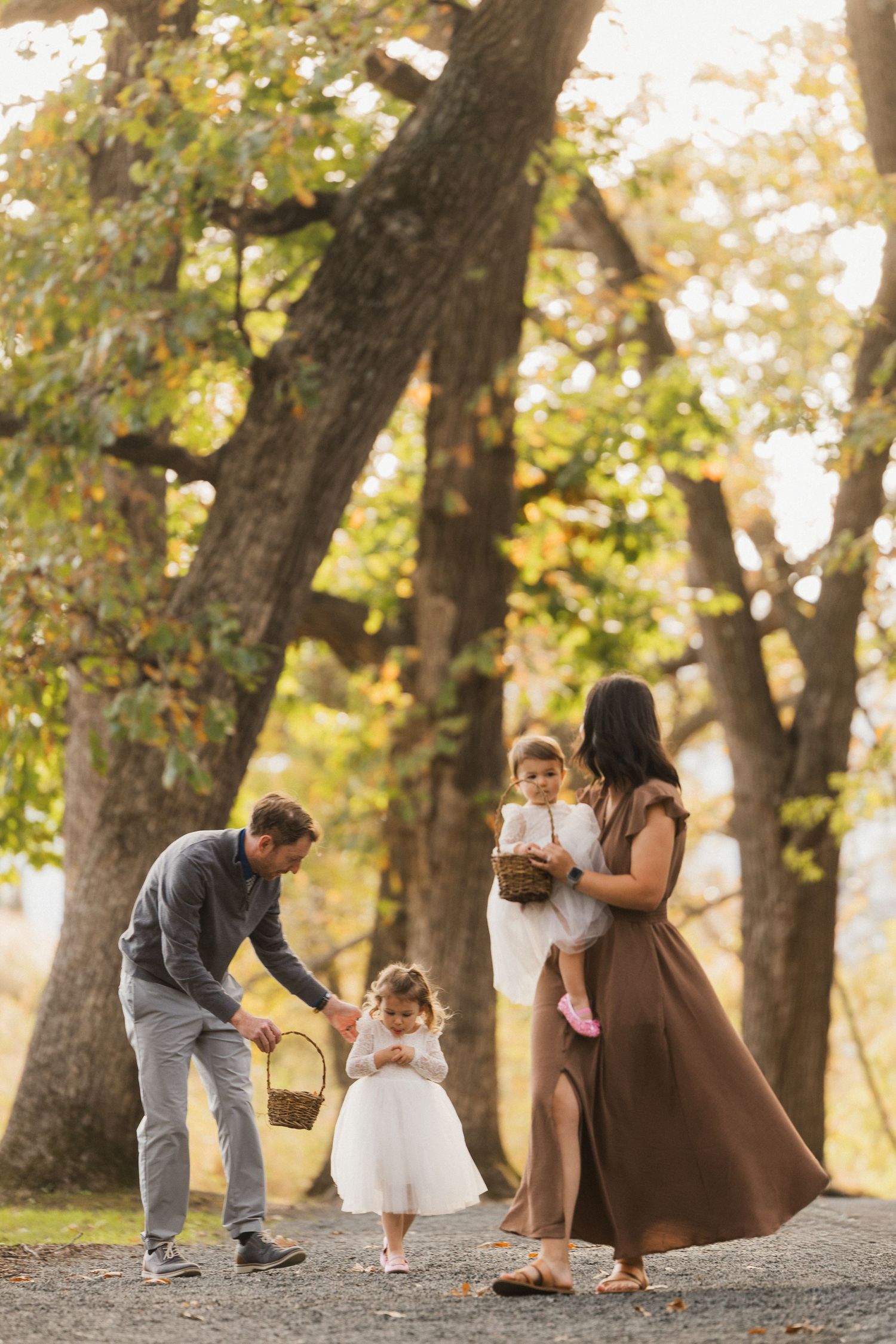 Family of four walks under tall autumn trees as two little girls in white dresses carry wicker baskets.