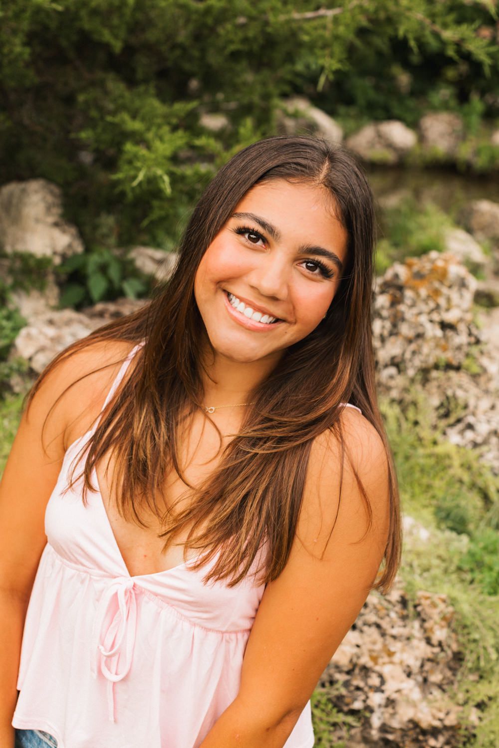 Portrait of someone in a pink camisole top smiling warmly against a natural rocky background.