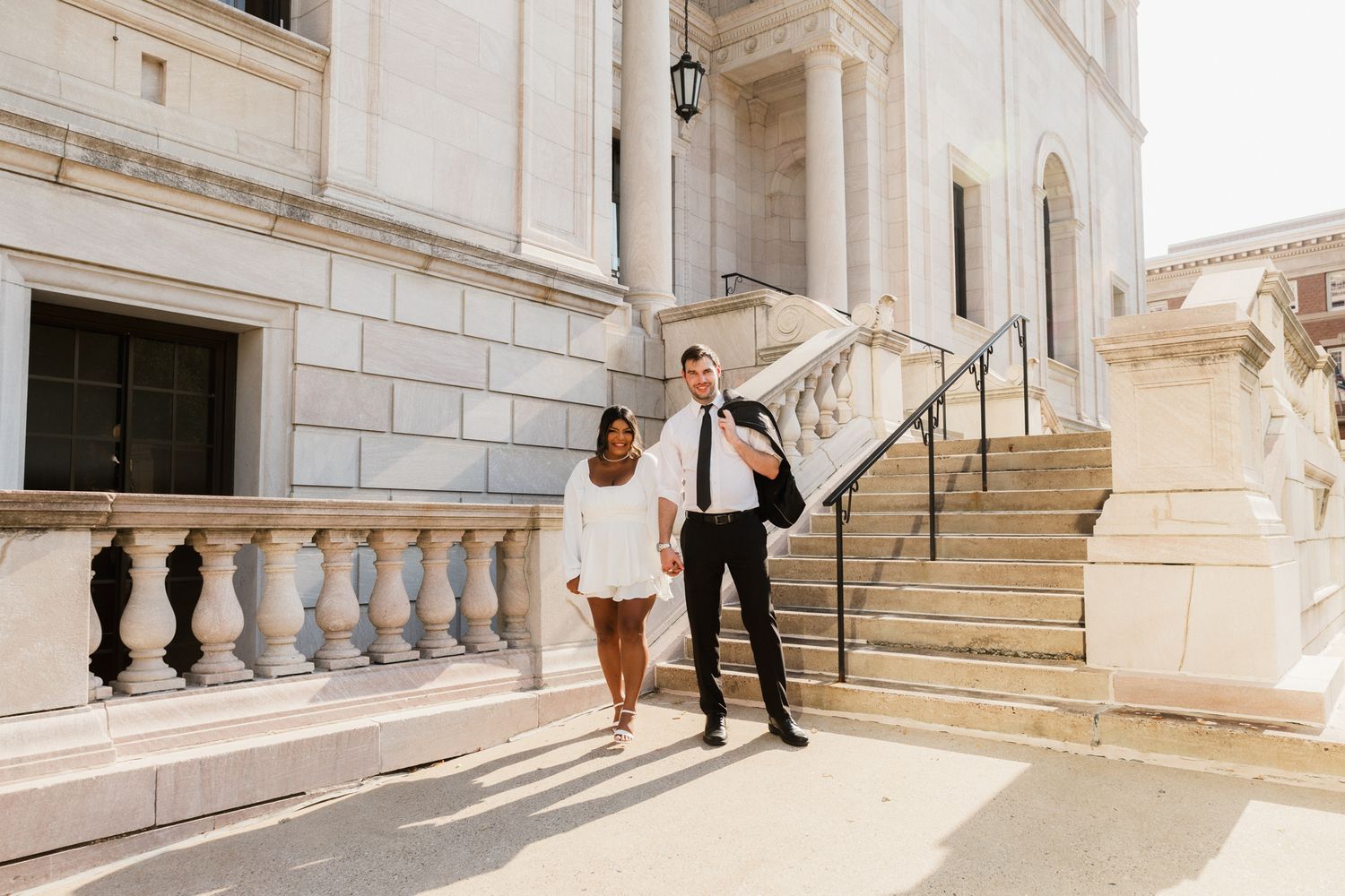 Couple in wedding attire walking hand-in-hand outside a grand white stone building with elegant balustrades.