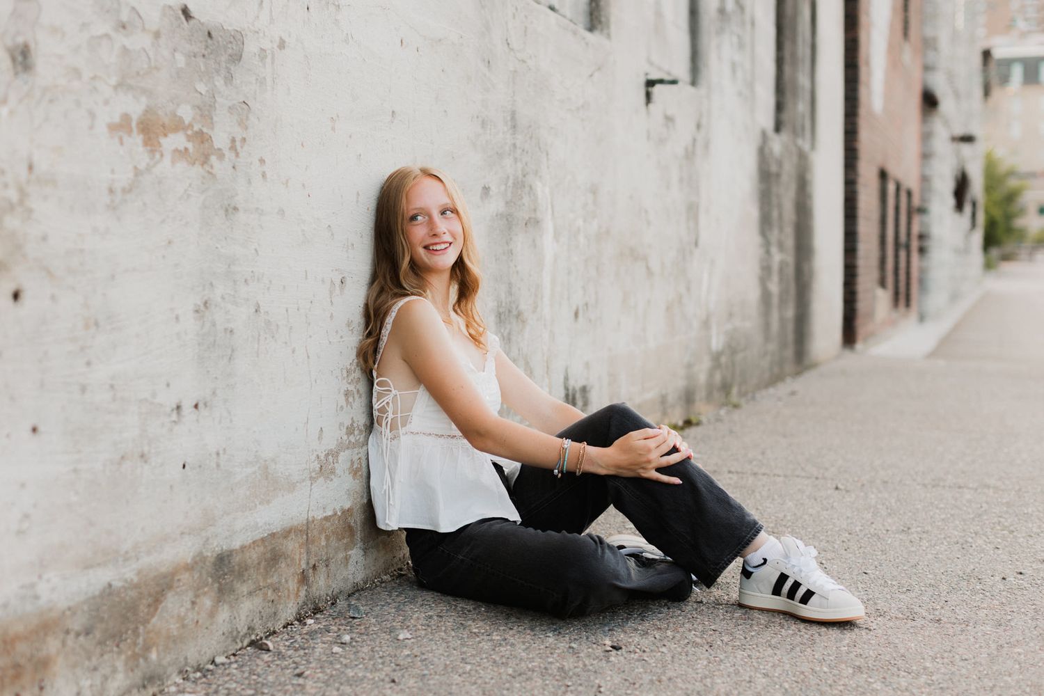 Smiling teenage girl in white top and black jeans sitting against rustic concrete wall outdoors.