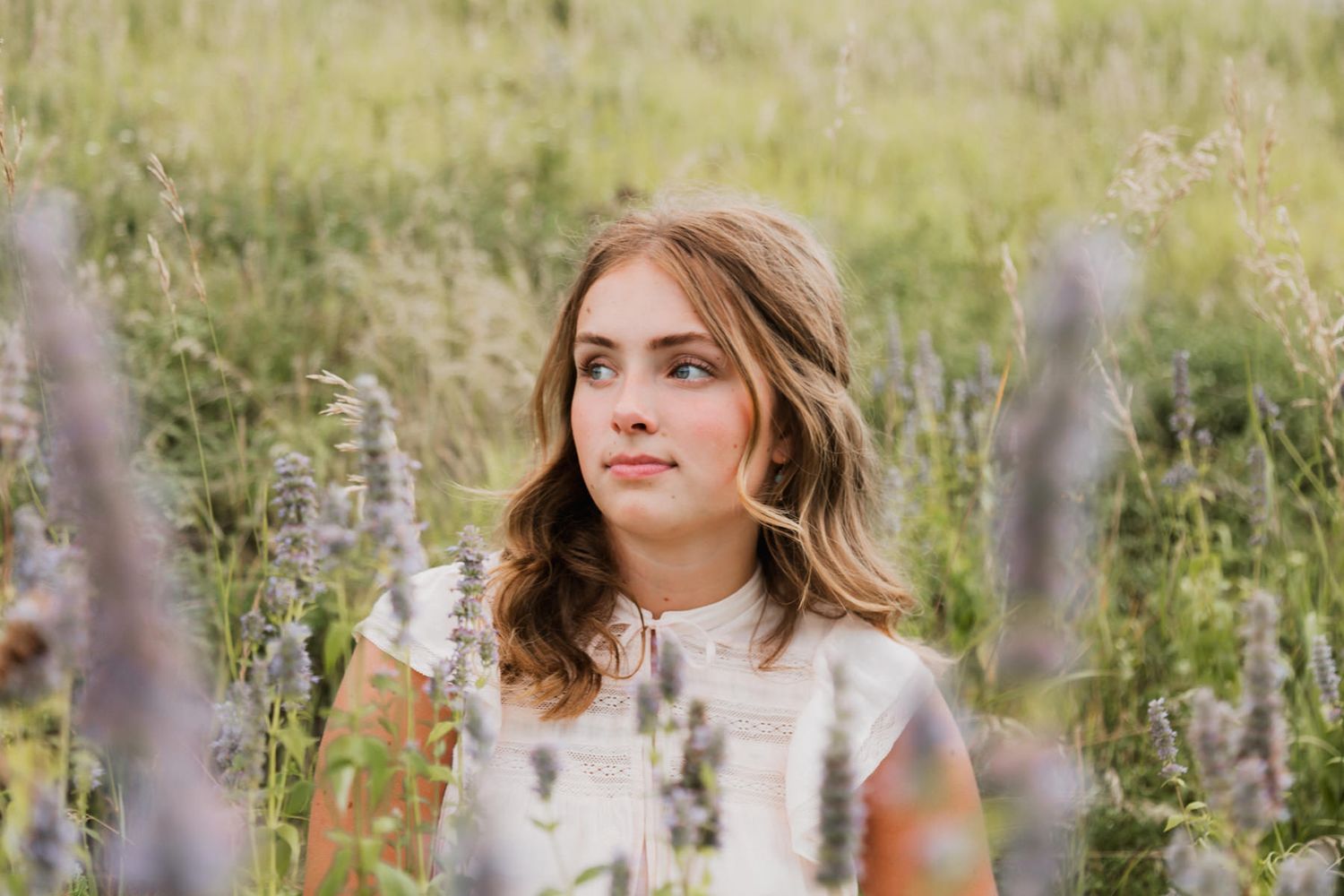 Young woman with wavy brown hair sitting peacefully among wildflowers in a meadow.