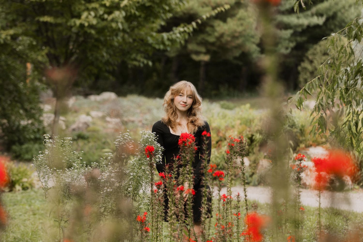 Young blonde woman standing among red poppies and greenery in a garden, wearing a black outfit.