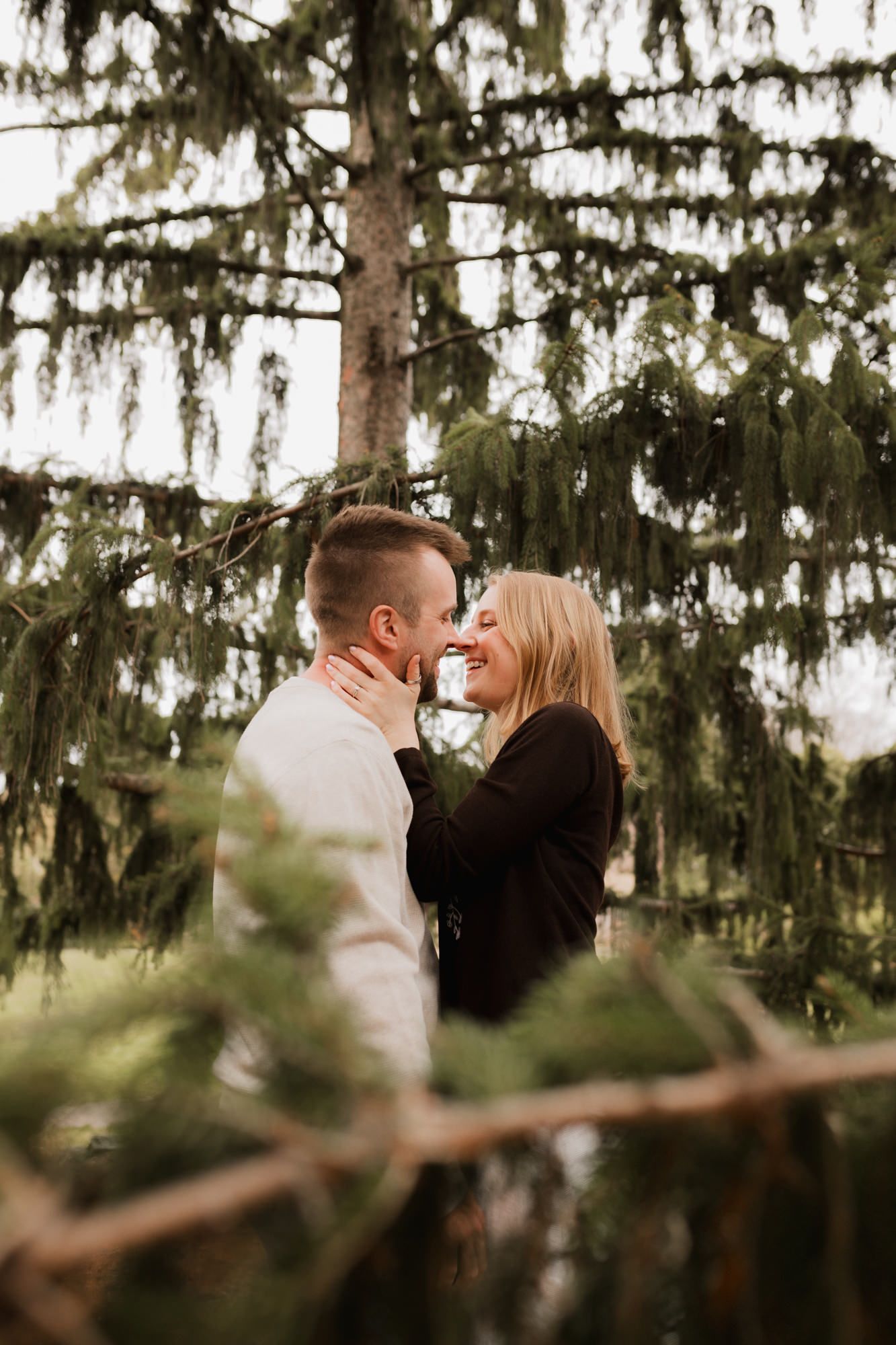 A couple shares a romantic moment nose-to-nose surrounded by lush green pine trees outdoors.