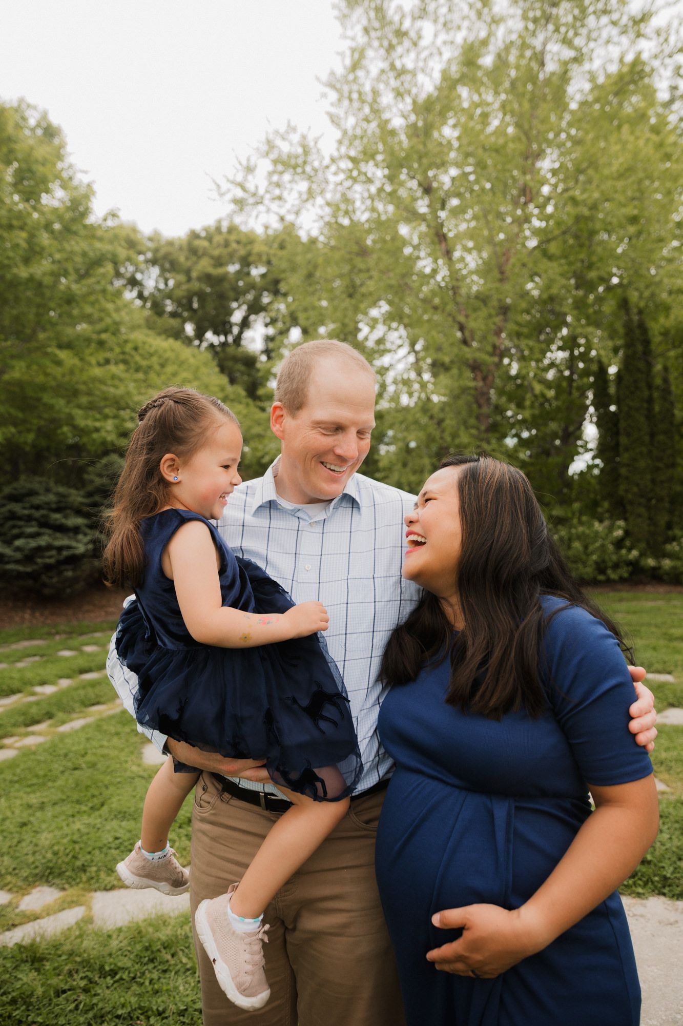 A happy family of three poses outdoors, with a man holding a toddler girl while a pregnant woman smiles beside them.