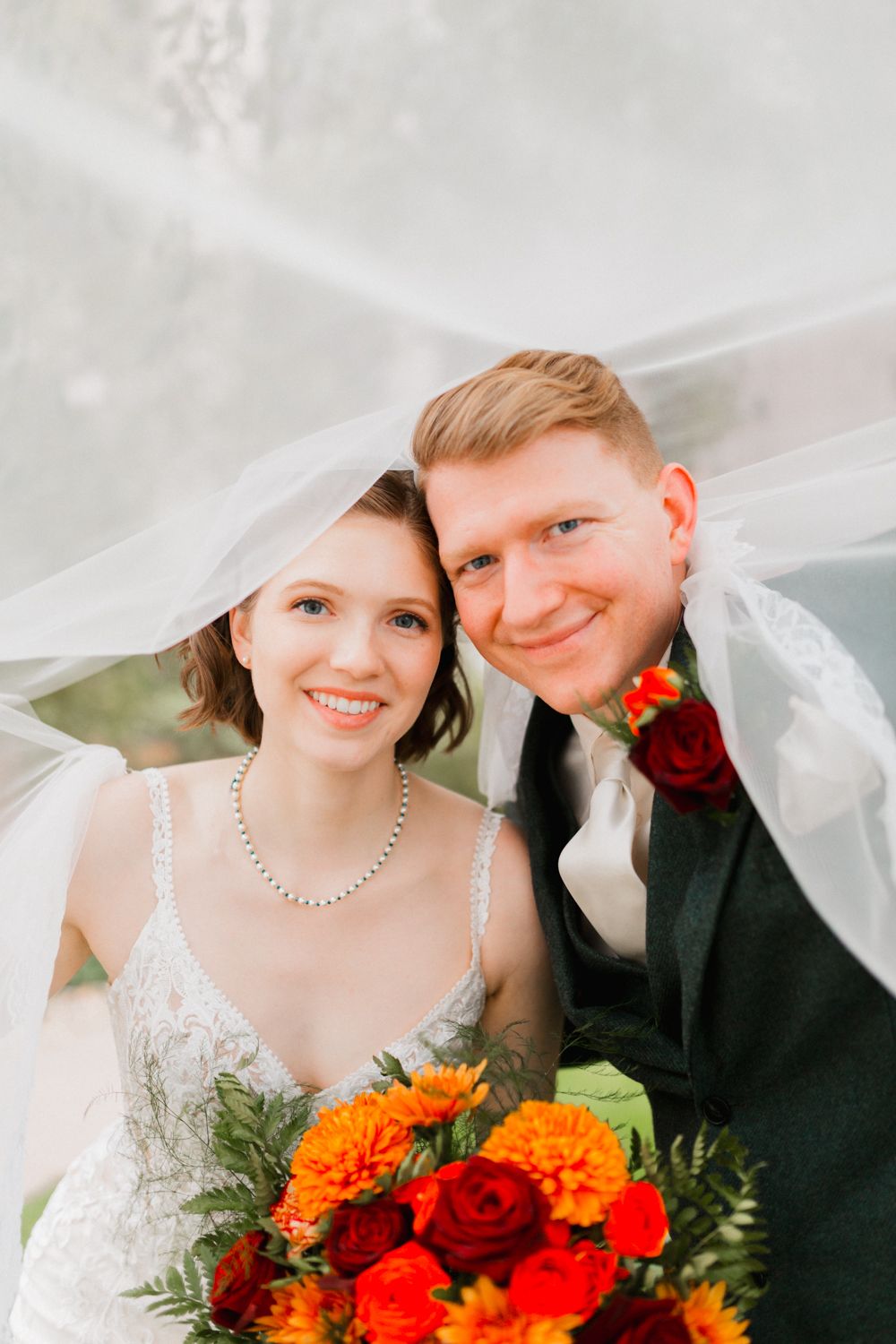 A wedding couple poses under a white veil with vibrant orange and red flowers in their bouquet.